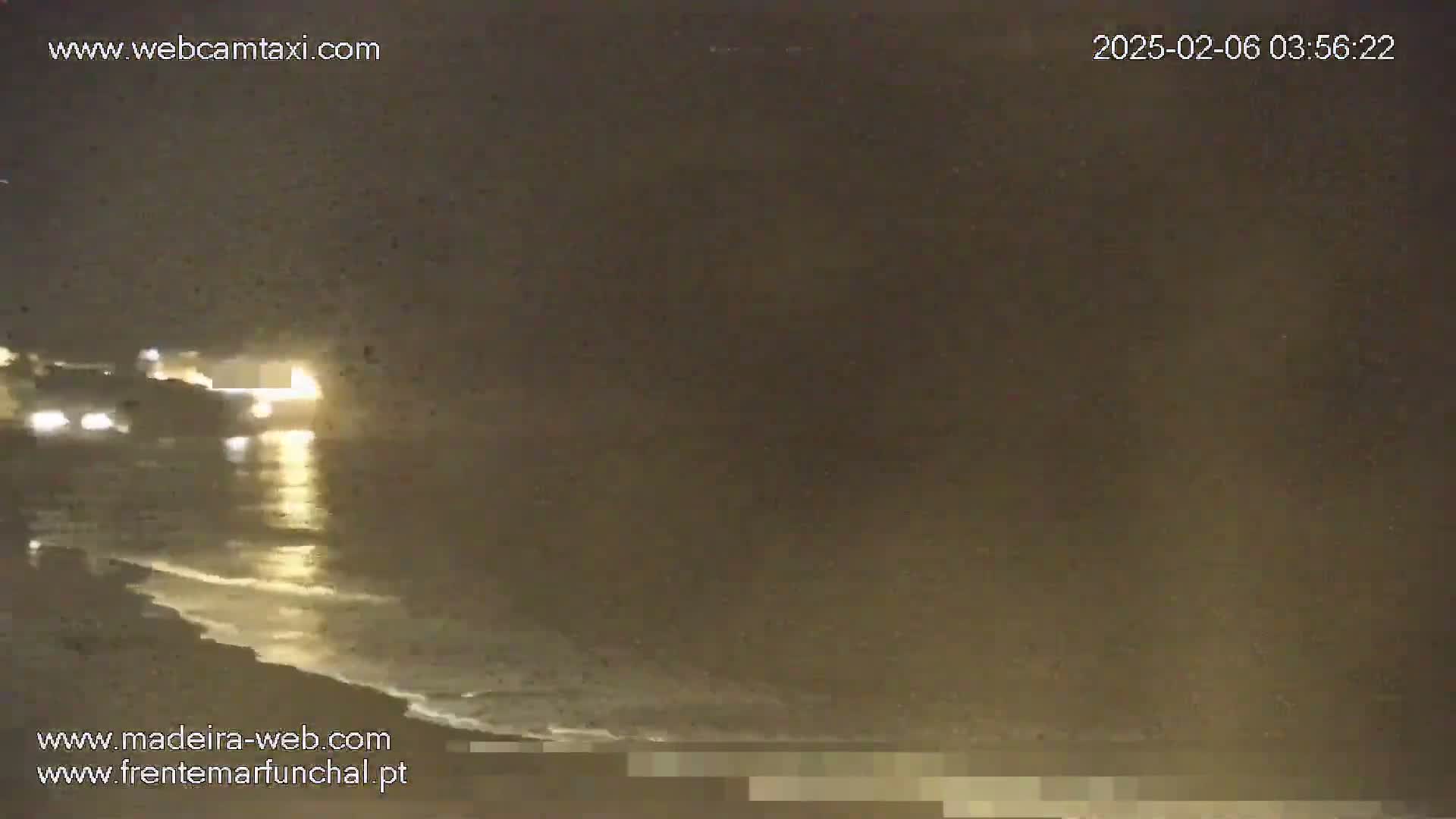A nighttime, dimly lit beach scene shows a dark ocean with small waves gently lapping the shore near some vehicles with headlights on.