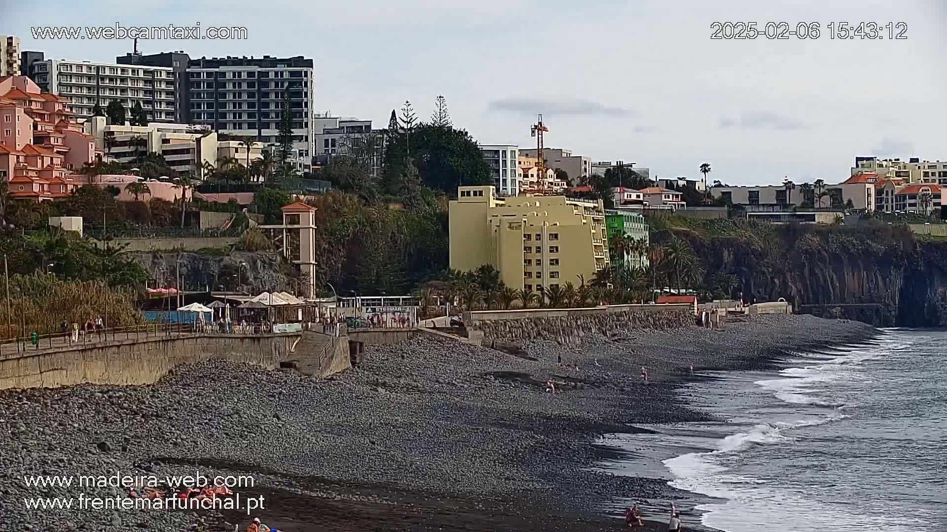 A dark-pebbled beach with gentle waves laps against the shore, under an overcast sky, beside a coastal town with several hotels and buildings built into a hillside.