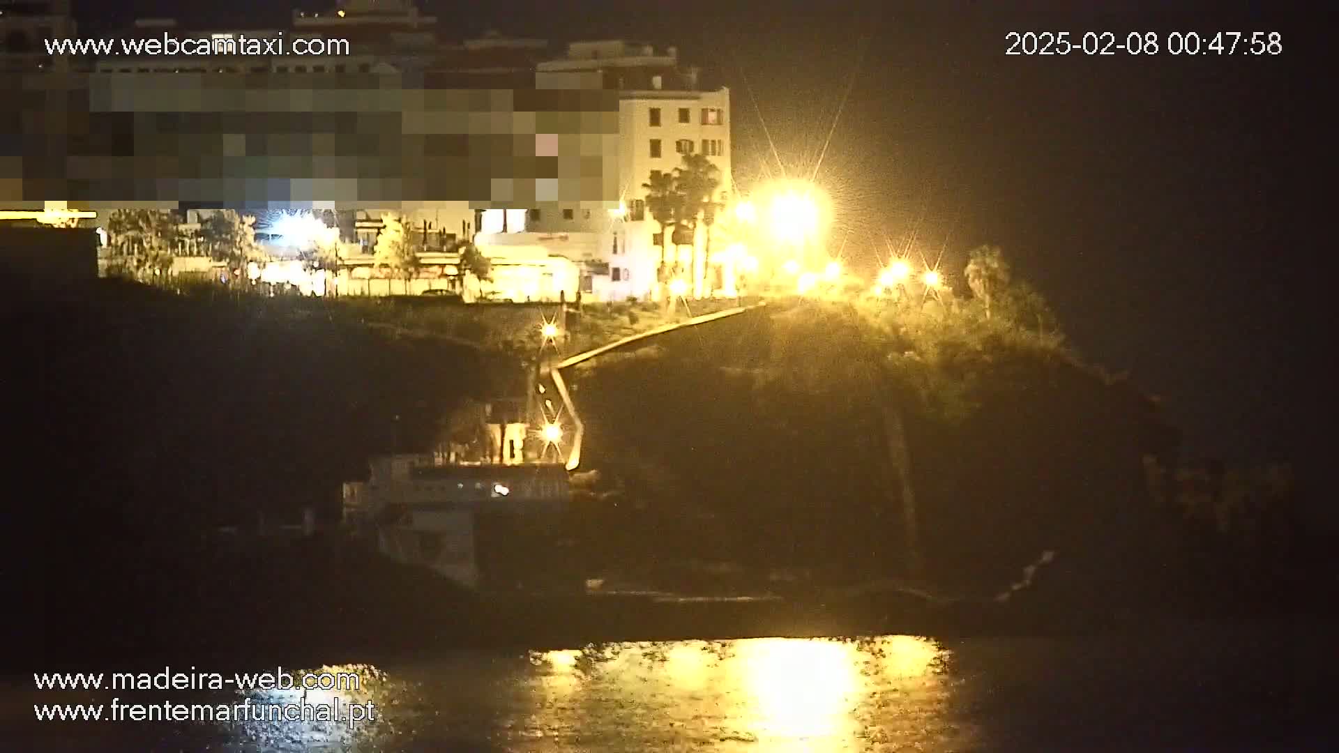 A nighttime view of a coastline with buildings and streetlights illuminating a dark, hilly landscape and calm water reflecting the lights.