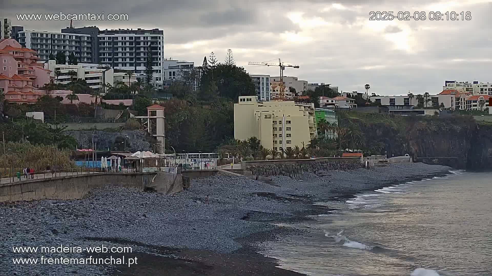 A rocky beach with gentle waves under an overcast sky, backed by buildings and a construction crane.