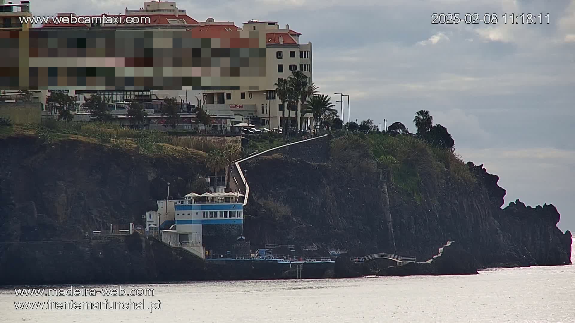 A multi-story building sits atop dark volcanic cliffs overlooking a calm body of water under a cloudy sky.