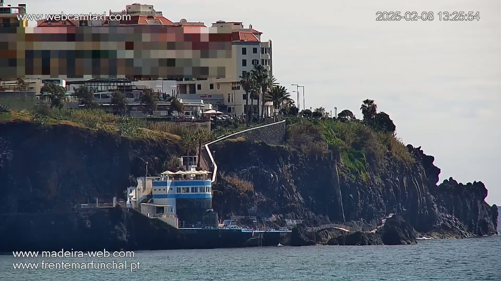 A coastal scene on a partly cloudy day shows dark volcanic rock cliffs with a building on the water's edge, a walkway, and a multi-story building on top of the cliff.