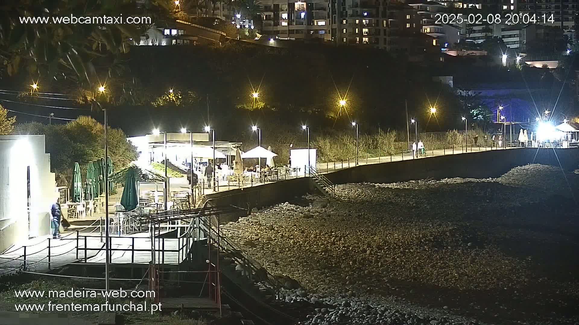 A nighttime view of an outdoor pebble beach area with a boardwalk, several lights, and outdoor seating at a restaurant under some umbrellas.