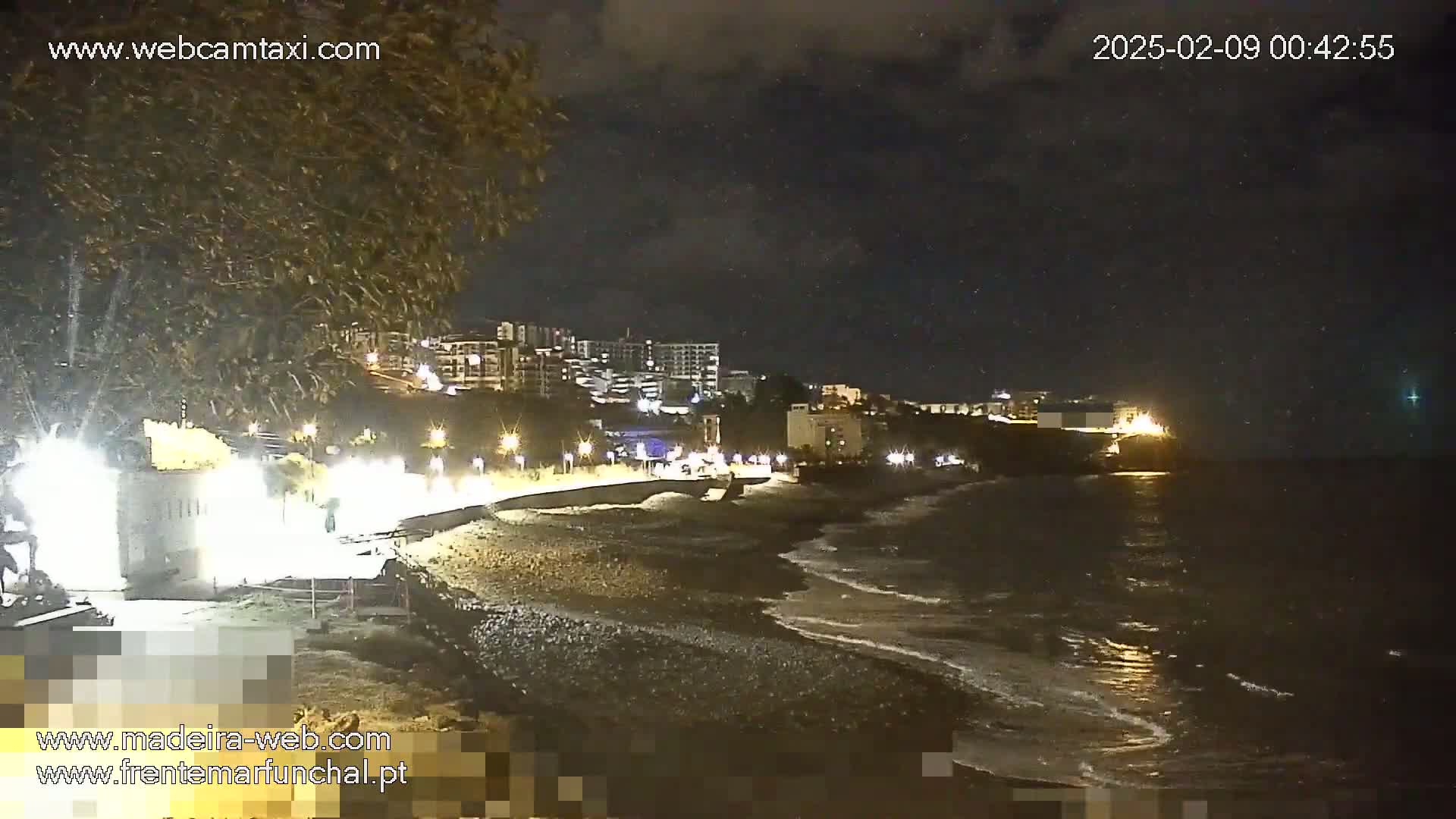 A nighttime beach scene shows a dark ocean with small waves lapping at a dark-colored, pebbled shore, under a partly cloudy night sky, with buildings and streetlights visible in the background.