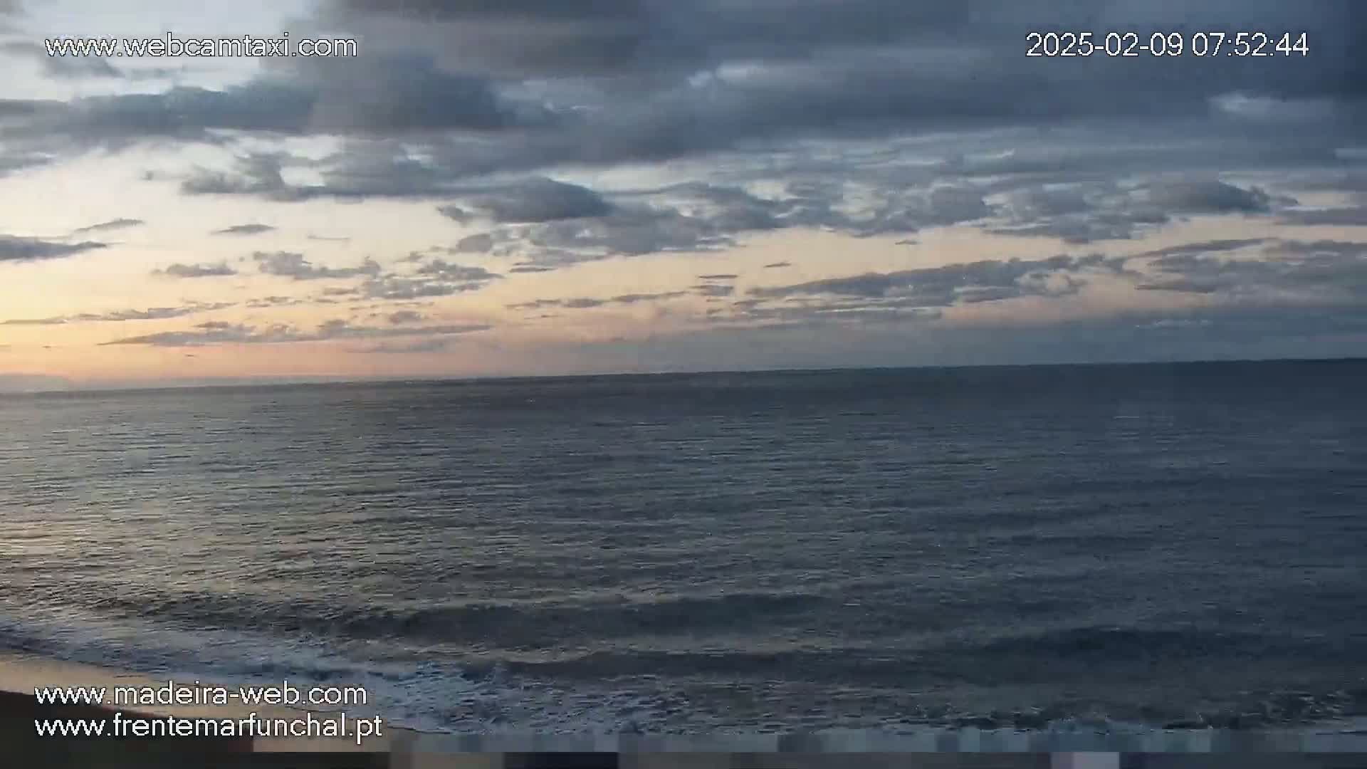 A partly cloudy sunrise over a calm ocean with small waves gently lapping the shore.