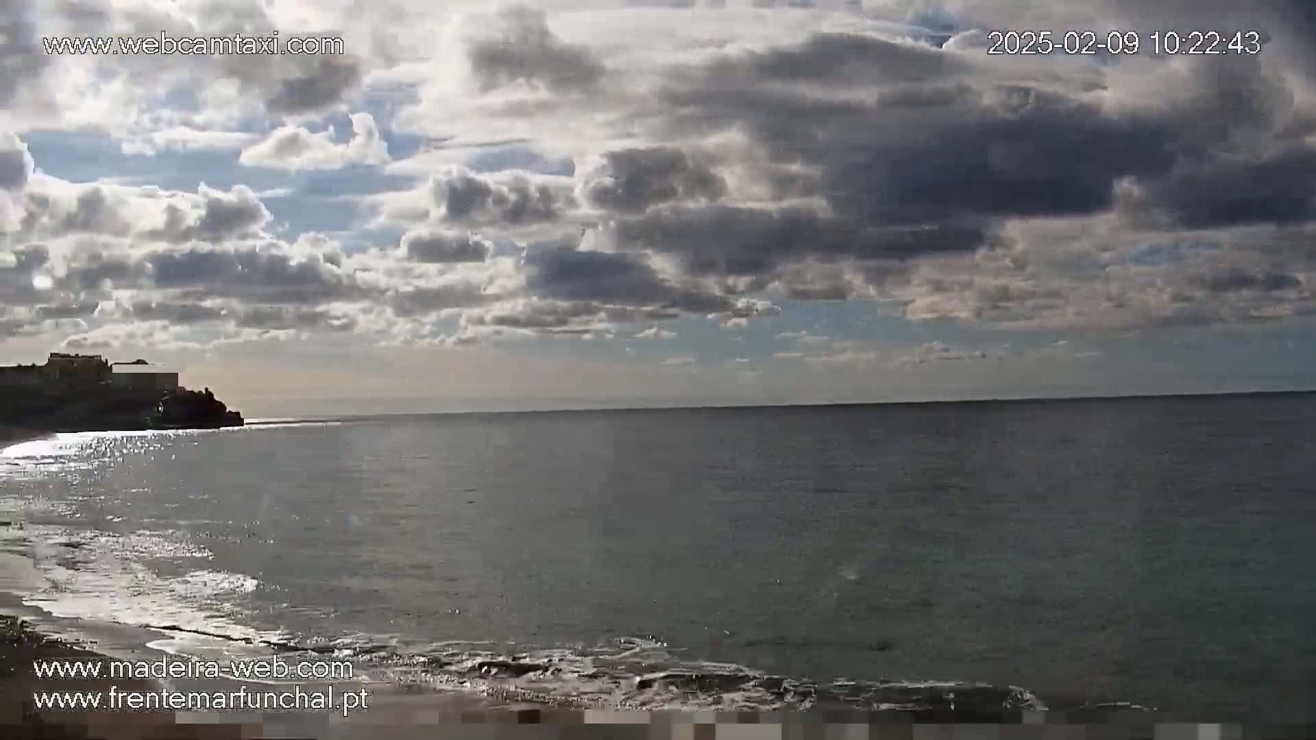 A mostly cloudy day reveals a calm ocean with a sandy beach and buildings visible on a distant cliff.