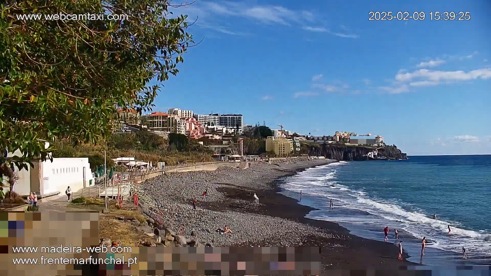 A dark-sand beach with a few people on it stretches along a coastline under a clear blue sky, backed by buildings and vegetation on a cliff.