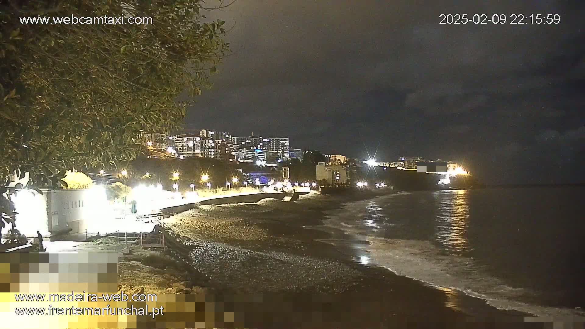 A nighttime view of a dark, pebble beach with gentle waves lapping the shore, under a partly cloudy sky, showing city lights and buildings in the background.