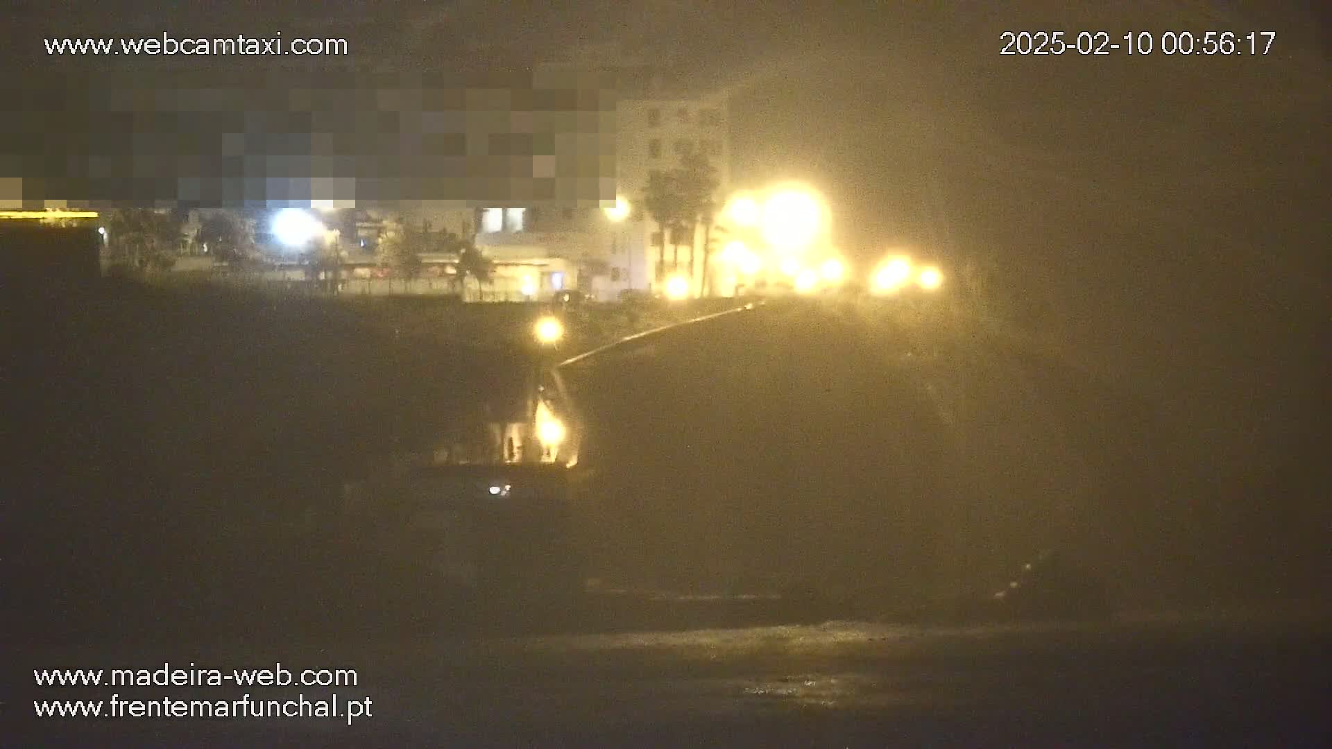A hazy night scene shows a blurry view of buildings and streetlights along a waterfront, with the lights reflecting on the water.