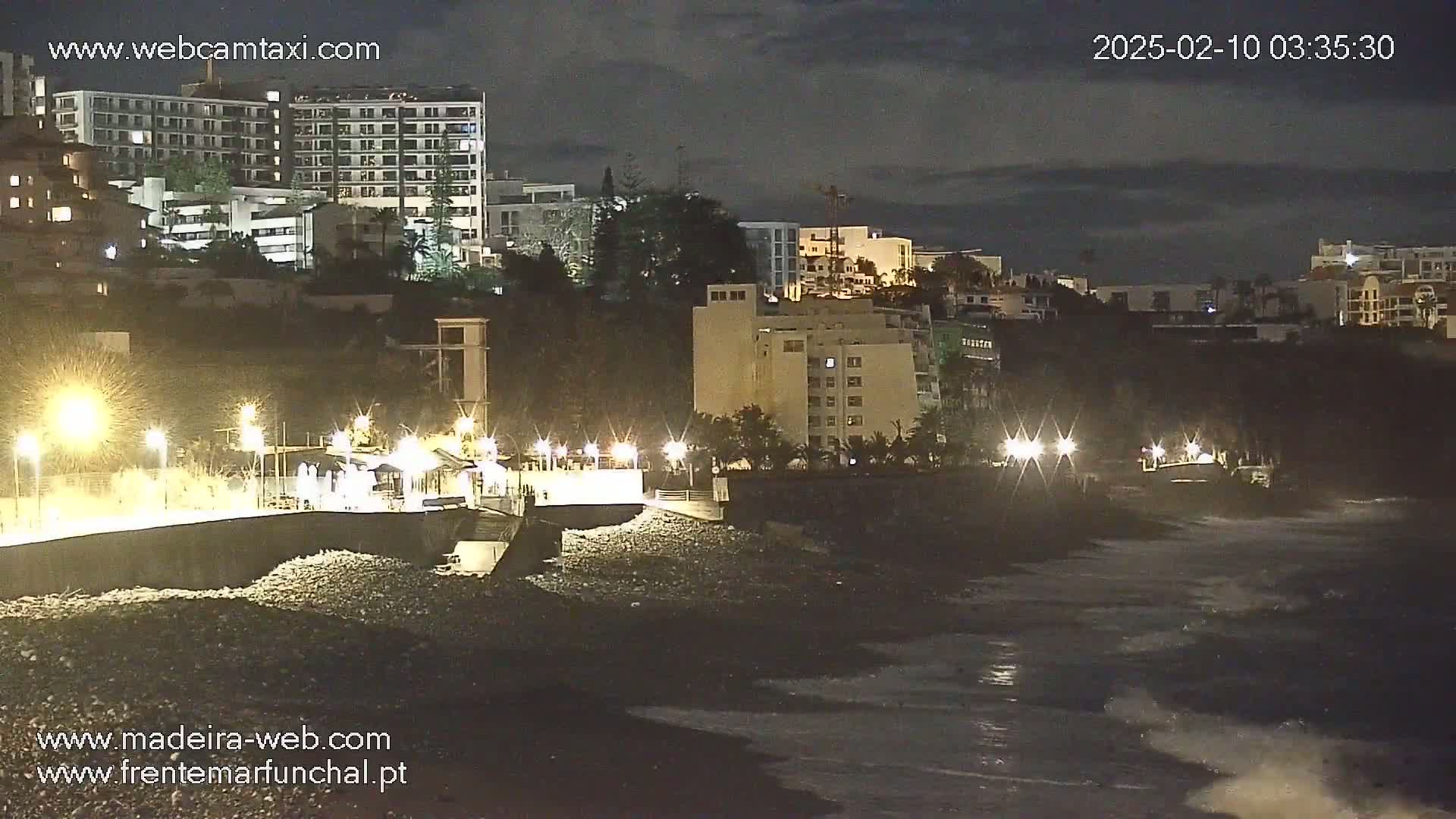 A nighttime coastal scene shows a dark, rocky beach with gentle waves lapping the shore, illuminated by streetlights, with multi-story buildings and vegetation on the hill above.