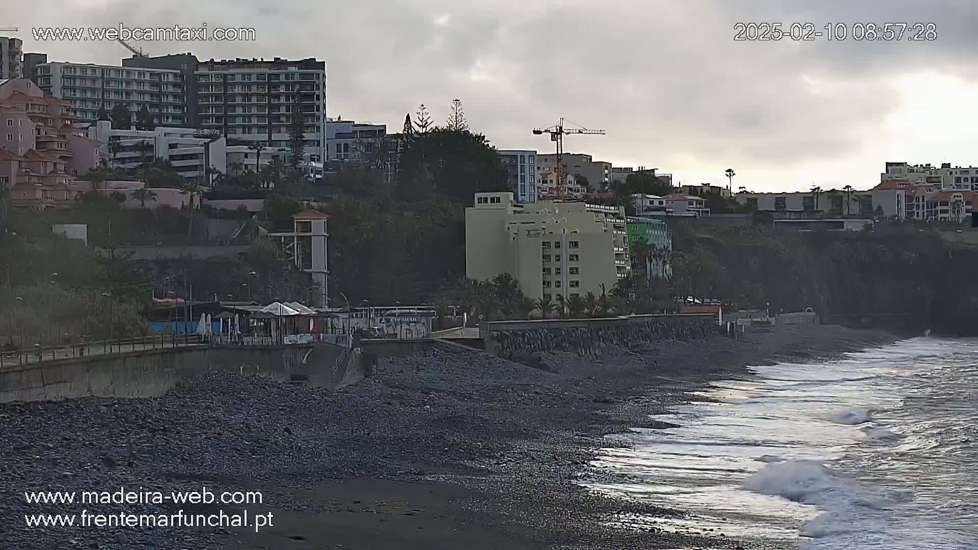 A dark, rocky beach with gentle waves laps against the shore under a cloudy sky, backed by a coastal town with multi-story buildings and construction cranes.