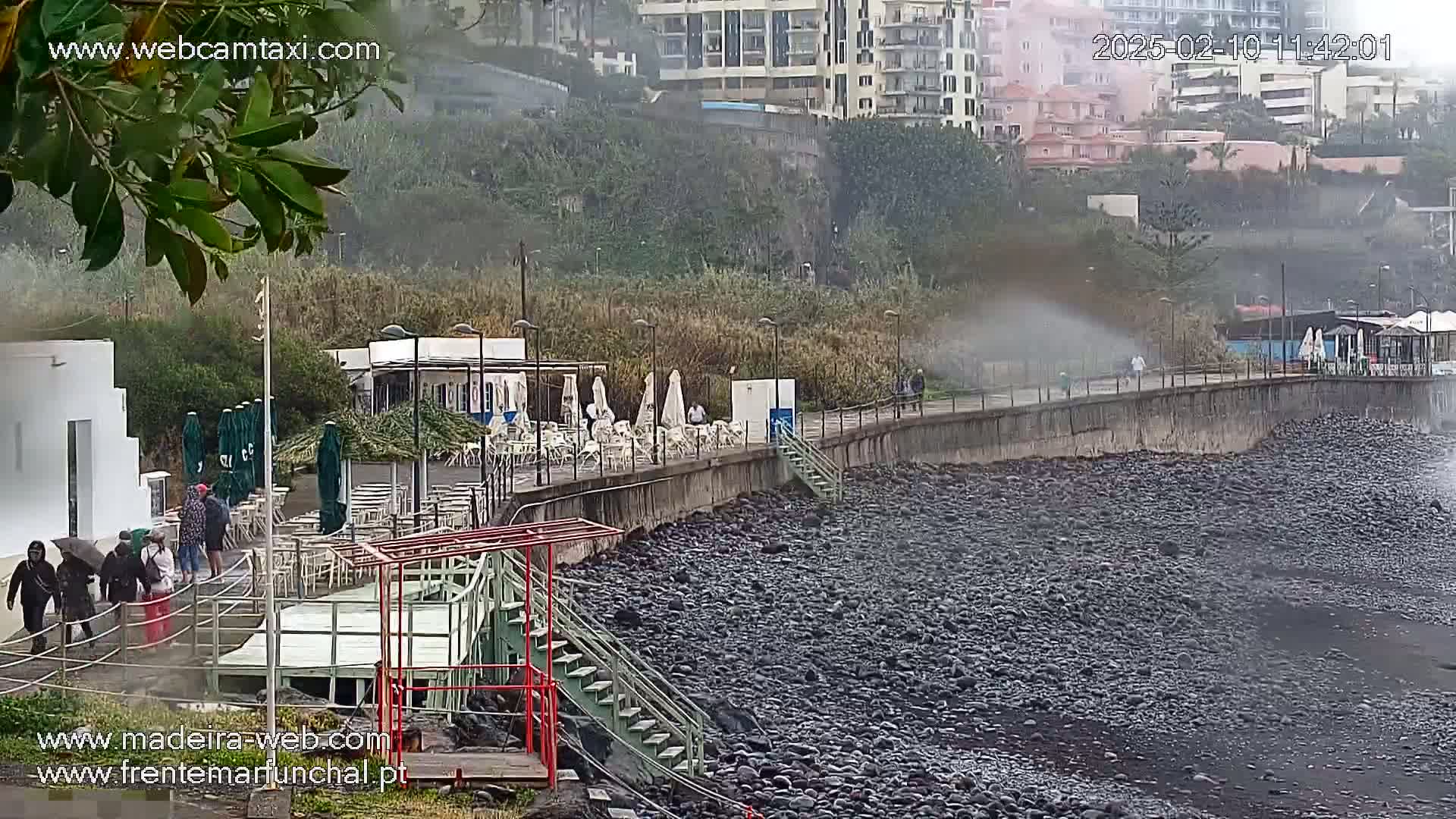 A rocky shoreline with an outdoor cafe and several people walking along a paved walkway in light misty conditions.