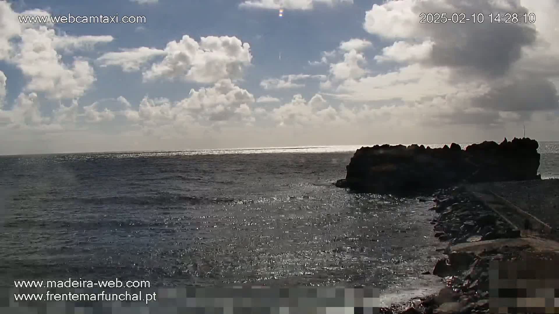 A partly sunny day shows a dark rocky outcrop extending into a somewhat choppy ocean under a mostly cloudy sky.