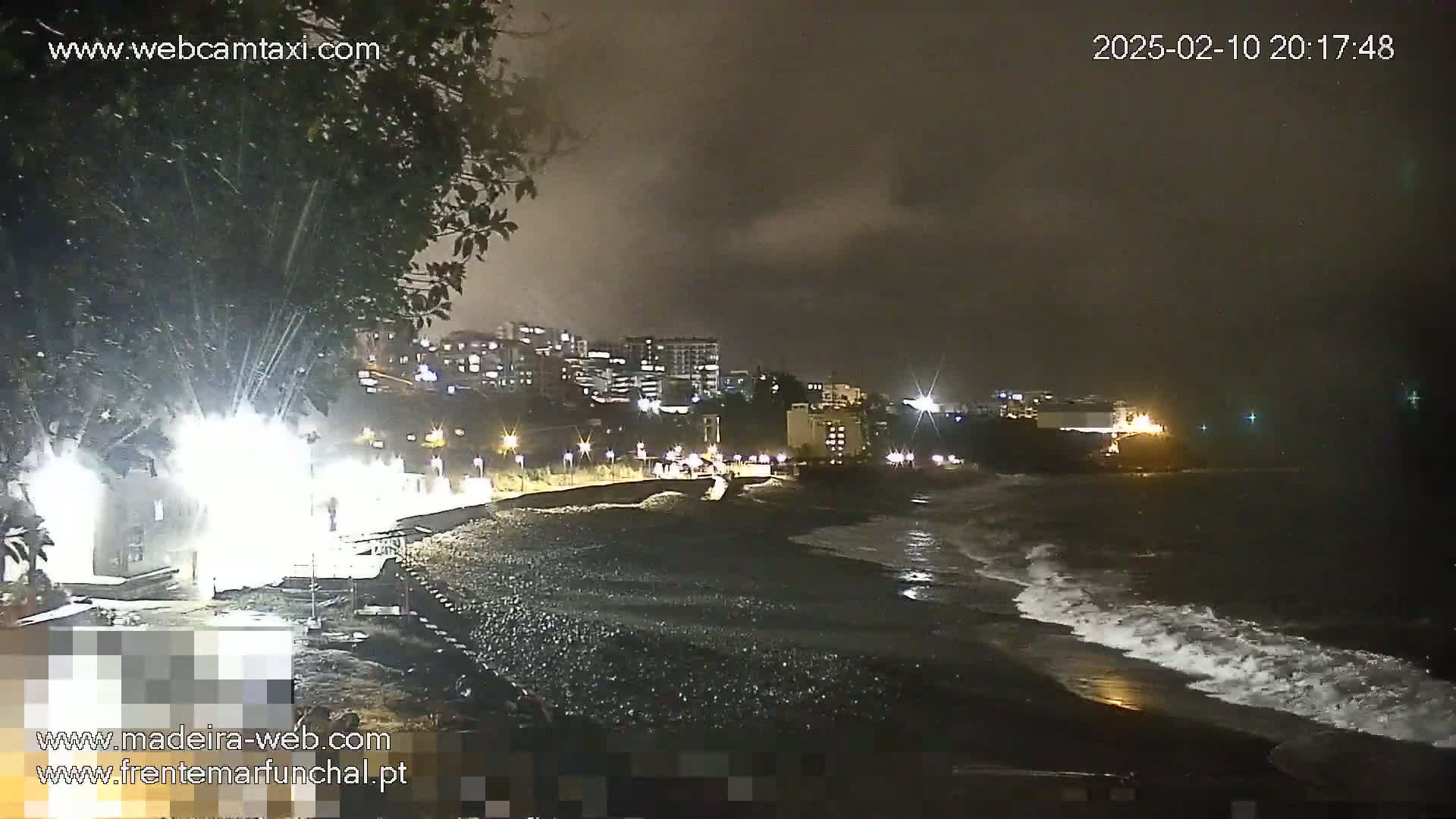 A nighttime view of a dark, pebble beach with gentle waves lapping the shore under a cloudy, dark sky,  lighted buildings are visible in the background.