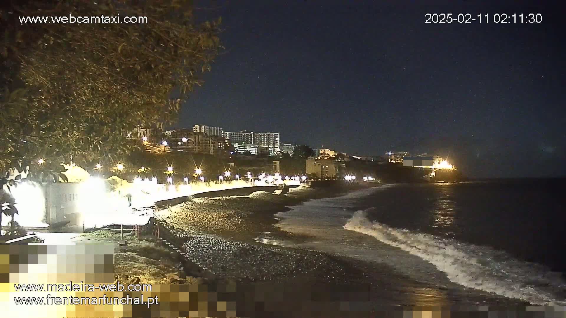 A nighttime view of a pebble beach with gentle waves lapping the shore, under a clear, starry sky, and buildings illuminated along the coast.