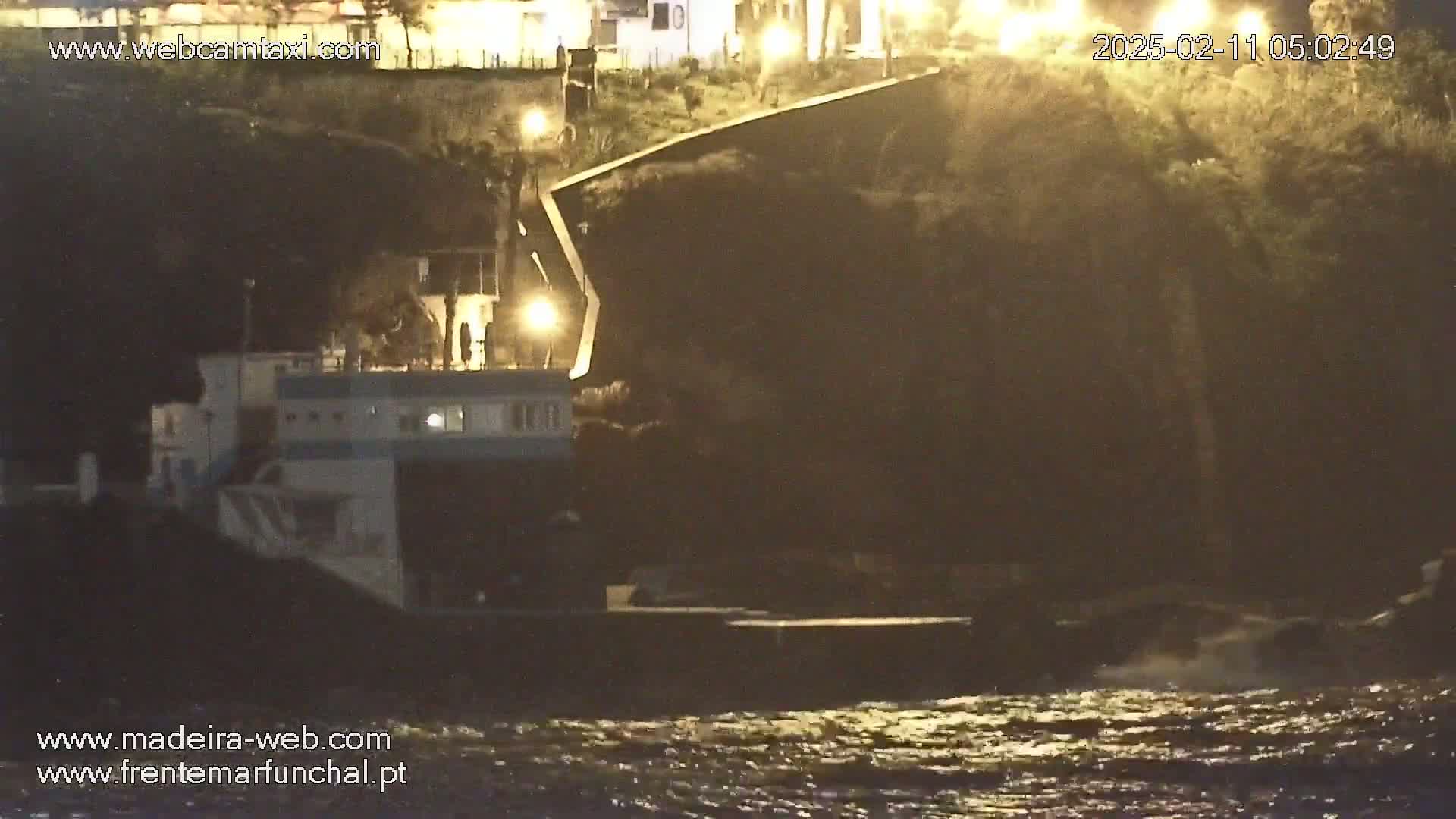 A boat is docked at night near a dark cliff face under somewhat choppy water.