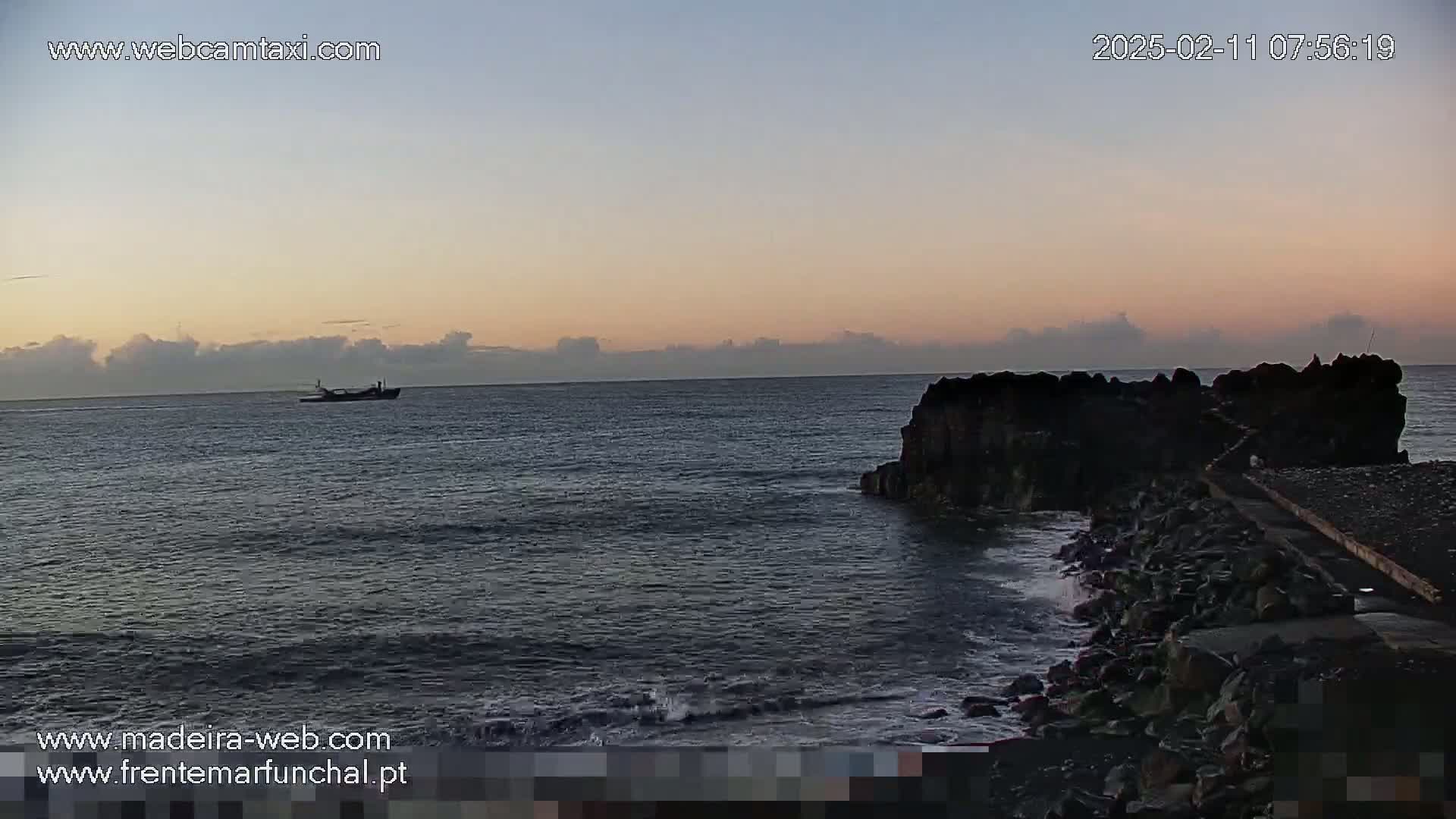 A calm ocean at sunrise shows a distant ship and dark rocky shoreline with a small wave breaking near the rocks.