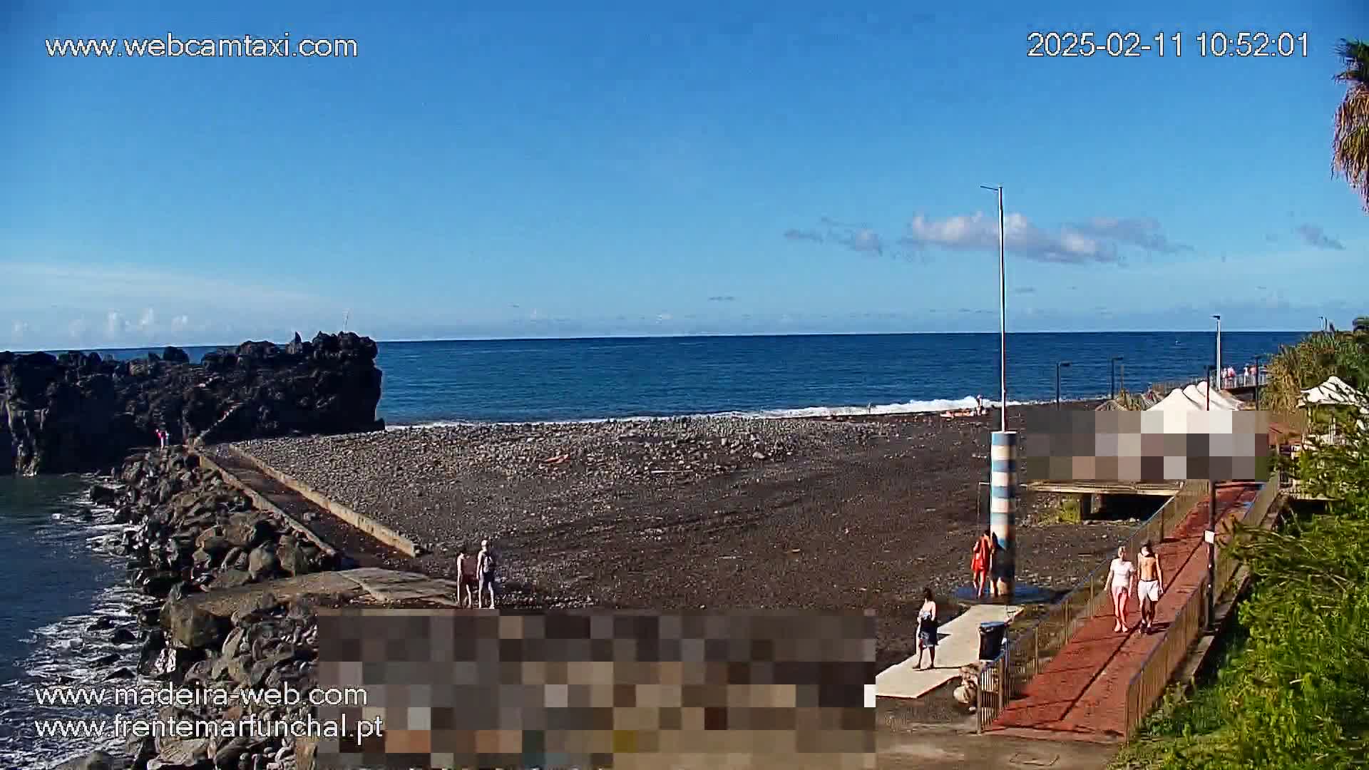 A dark-sand beach with a few people on it stretches from a rocky point into a calm blue ocean under a mostly sunny sky.