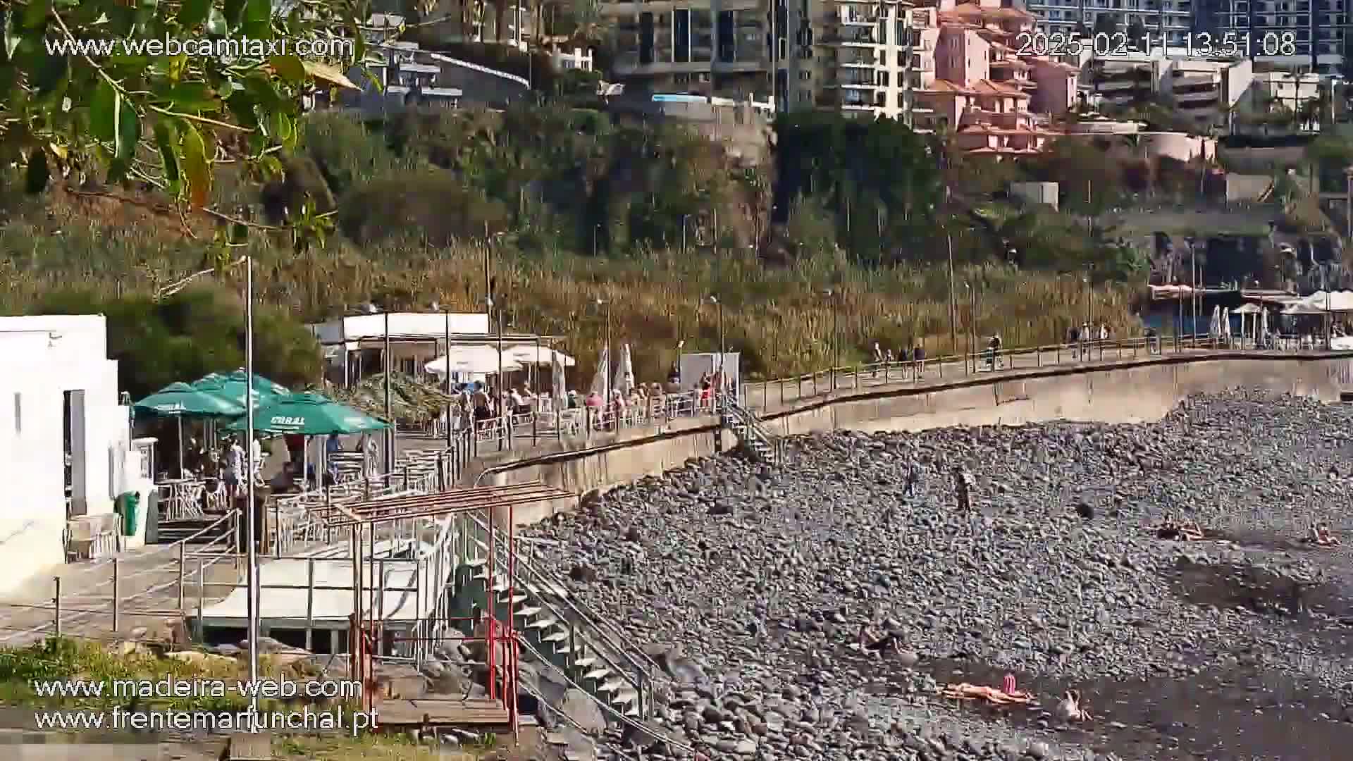 A sunny day at a rocky beach with people relaxing near a boardwalk and outdoor cafes.