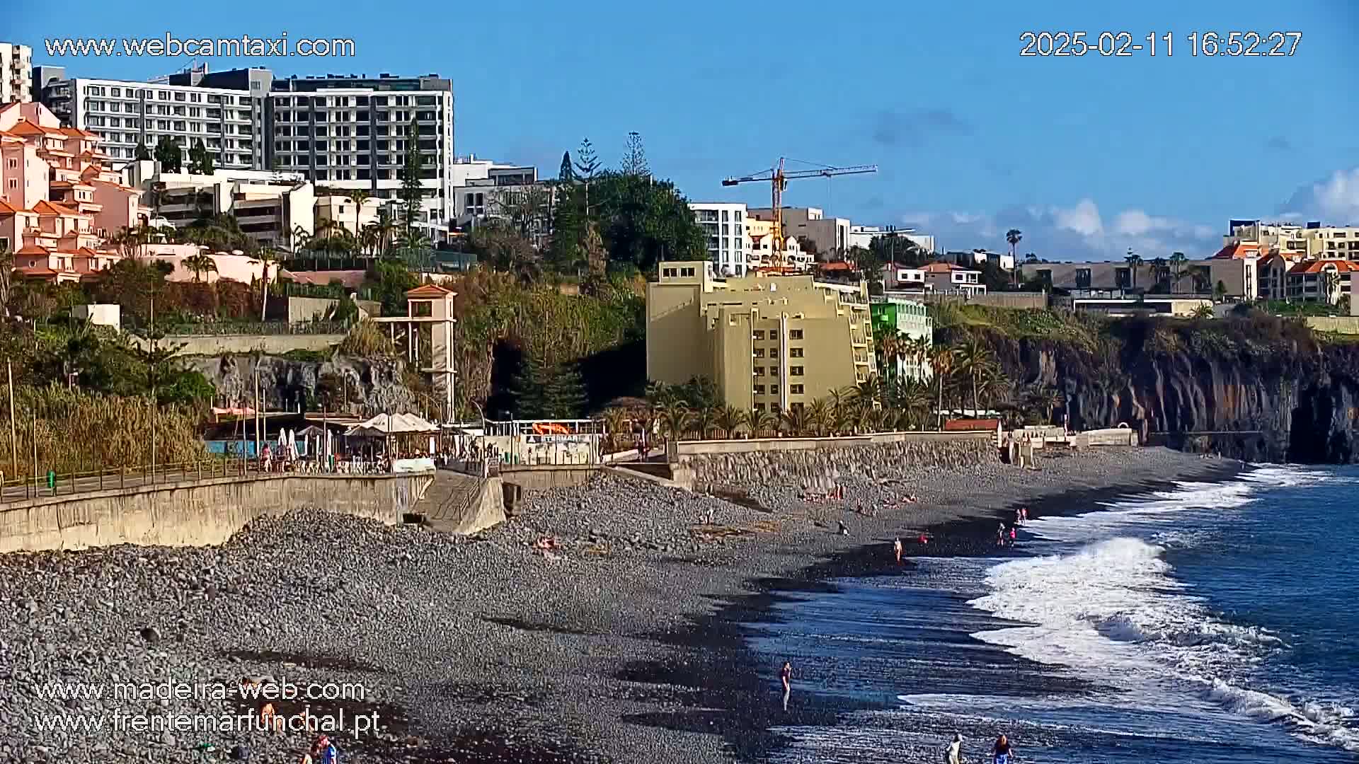 A mostly sunny day shows a rocky black-sand beach with small waves lapping the shore, in front of hotels and buildings situated on a cliffside.