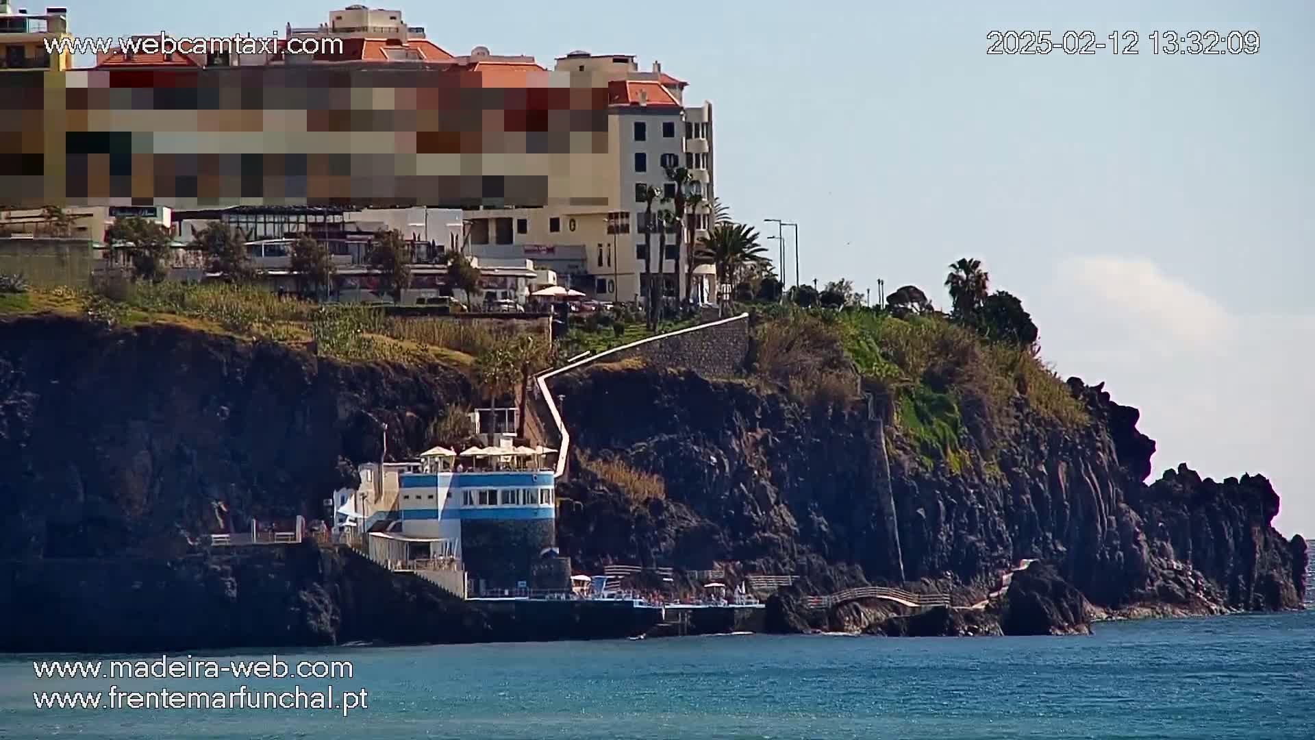 A coastal scene on a sunny day shows a multi-story building perched atop dark volcanic cliffs, with a smaller building and a walkway leading down to the ocean.