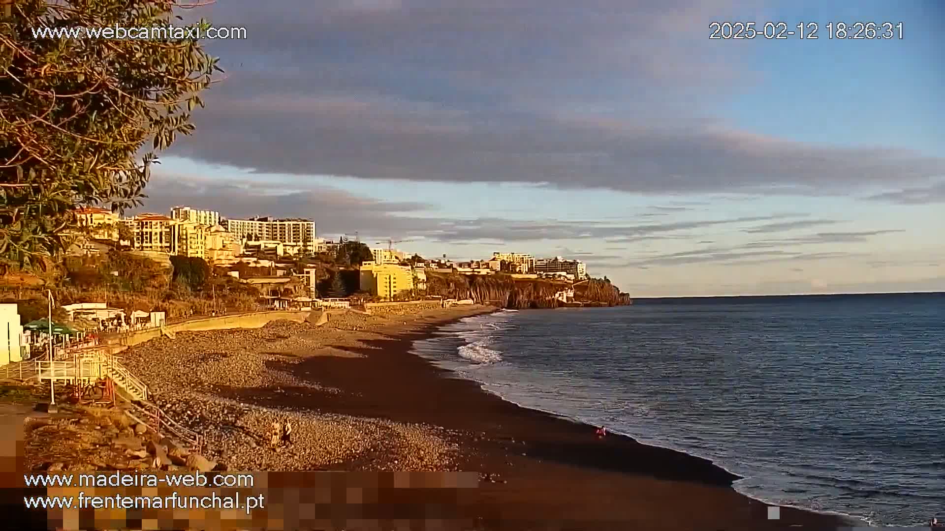 A dark-sand beach with gentle waves laps against the shore under a partly cloudy sky,  buildings are visible on the hillside above.