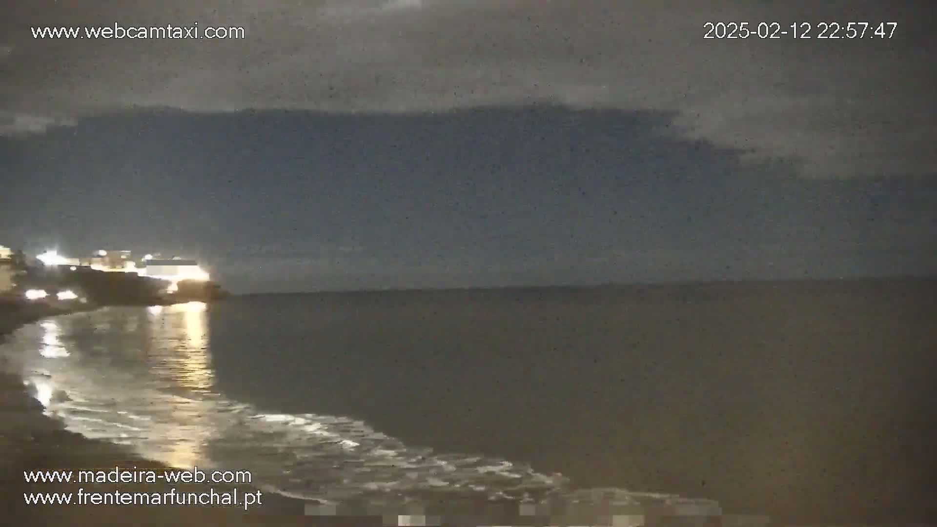 A nighttime coastal scene shows a dark ocean with gentle waves lapping a shoreline, illuminated by lights from buildings on the land.