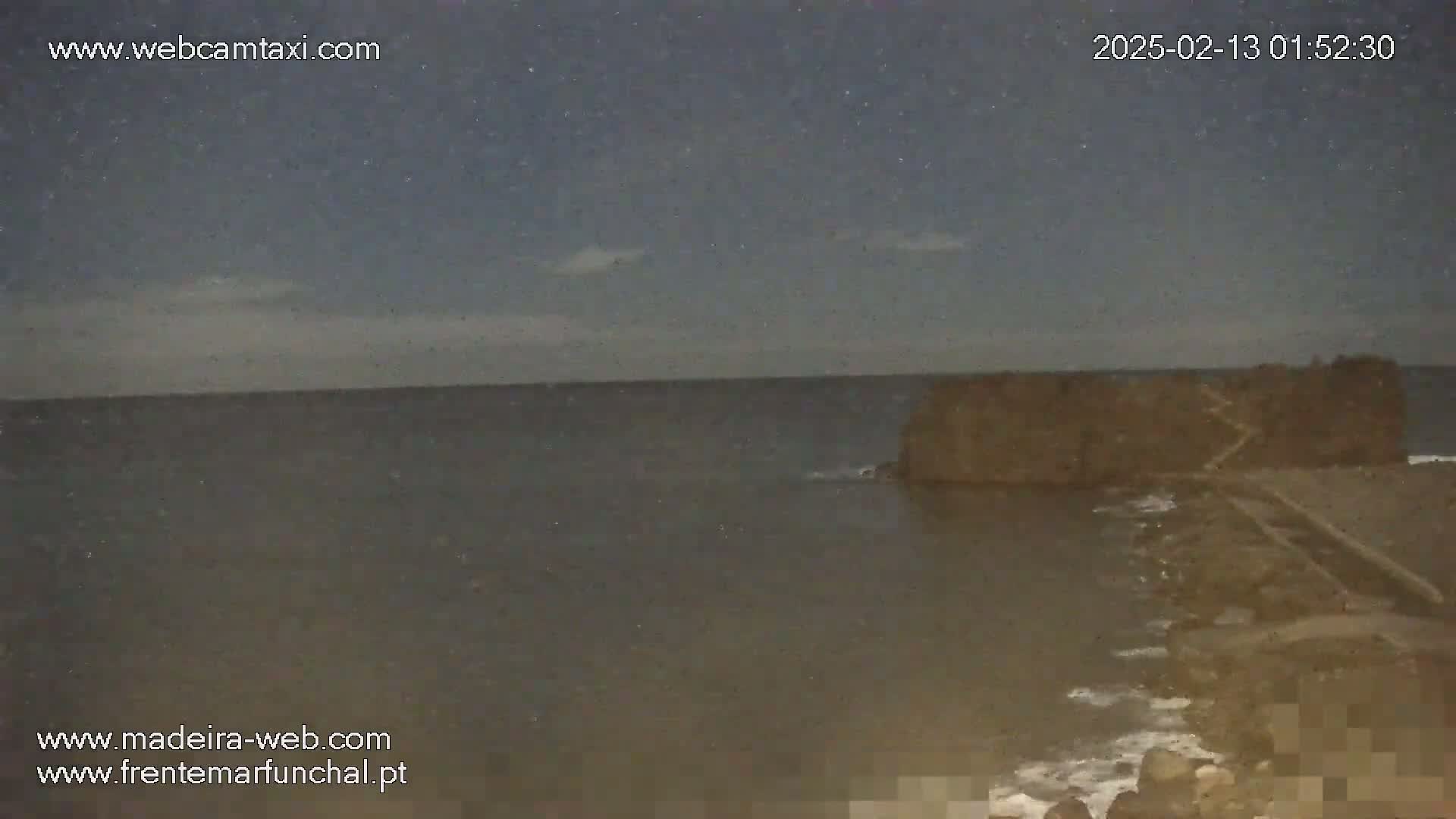 A dark, nighttime ocean scene shows a rocky outcropping connected to the shore by a path under a slightly cloudy sky.