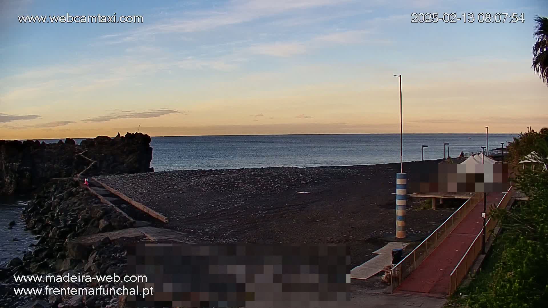 A dark-colored beach with a calm ocean under a mostly clear, twilight sky is shown, with a walkway and some structures visible near the water.