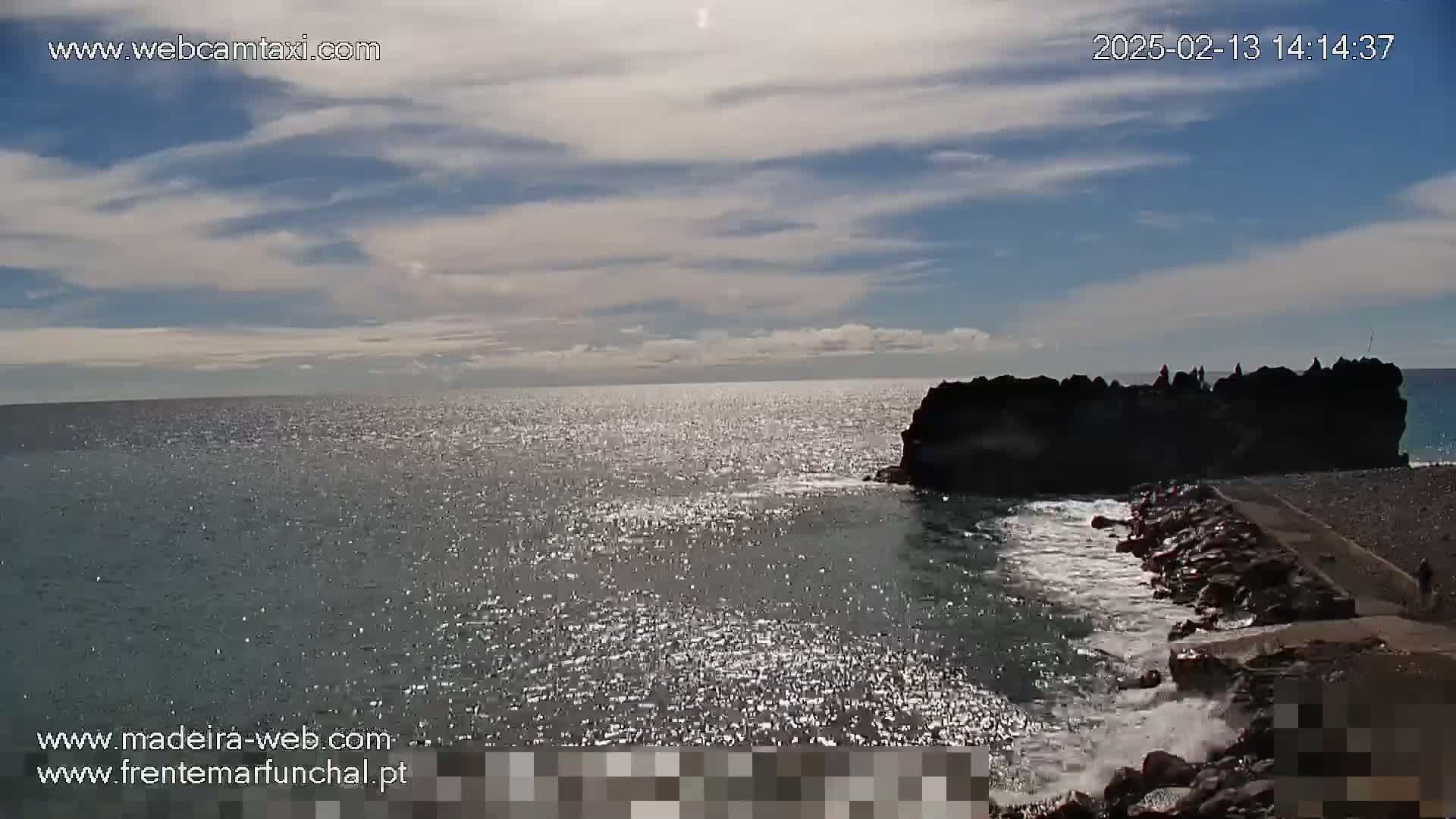 A sparkling ocean under a partly cloudy sky meets a dark rocky outcropping with a few people silhouetted on top.