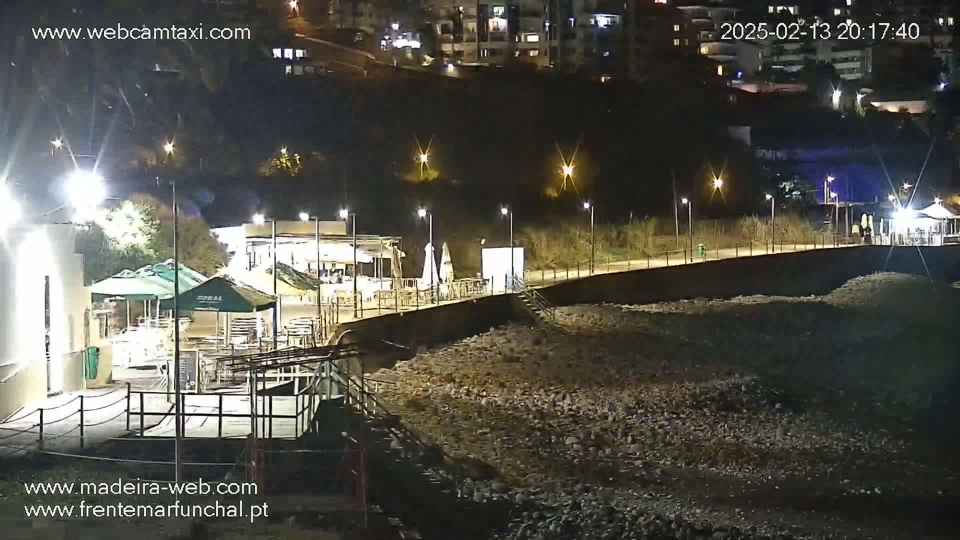 A nighttime view of a rocky beach area with illuminated buildings, walkways, and outdoor seating areas along a waterfront.