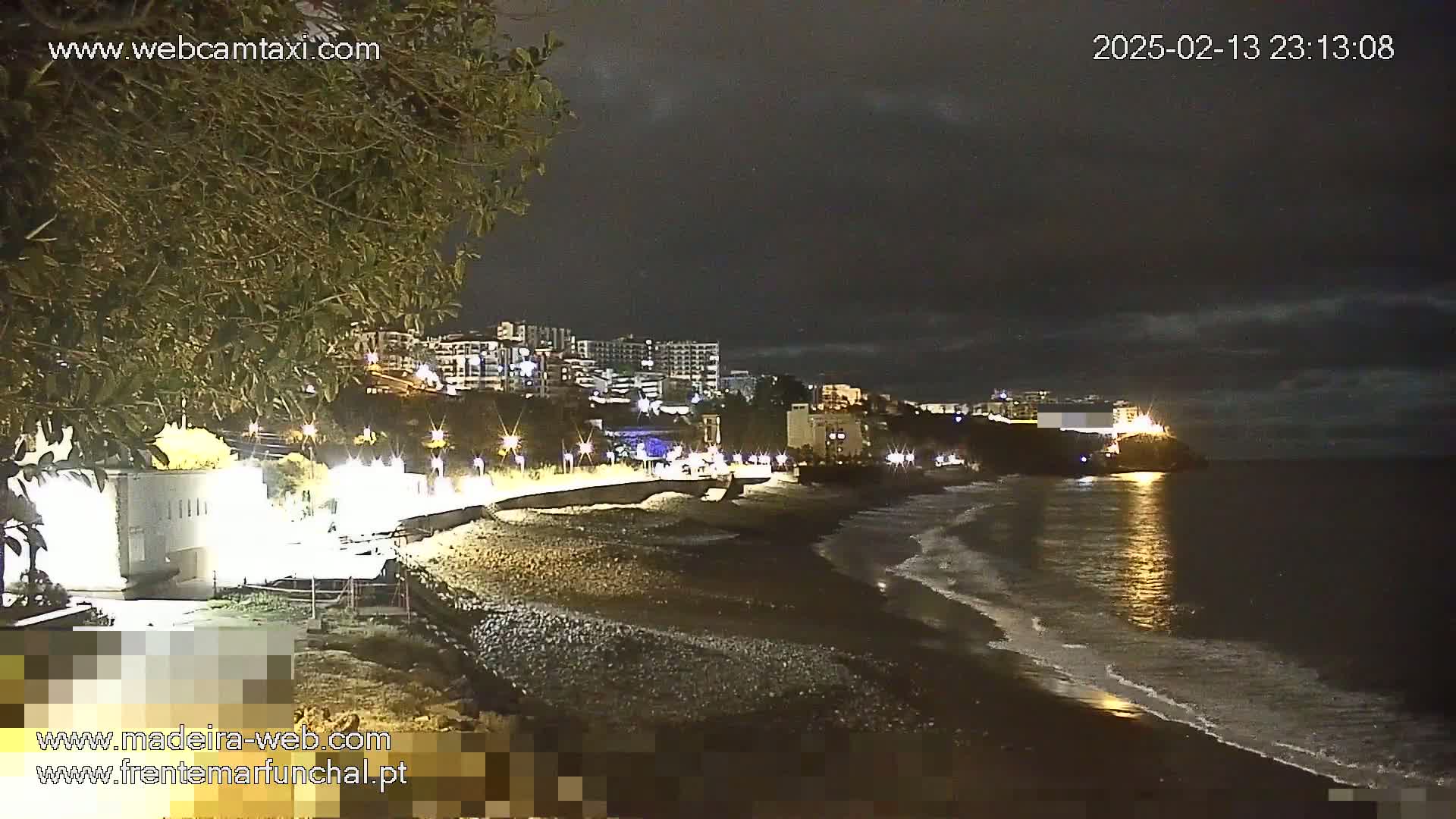 A dark, nighttime coastal scene shows a pebble beach with gentle waves lapping the shore, illuminated buildings on a hill overlooking the water, and a few lights along a waterfront path.