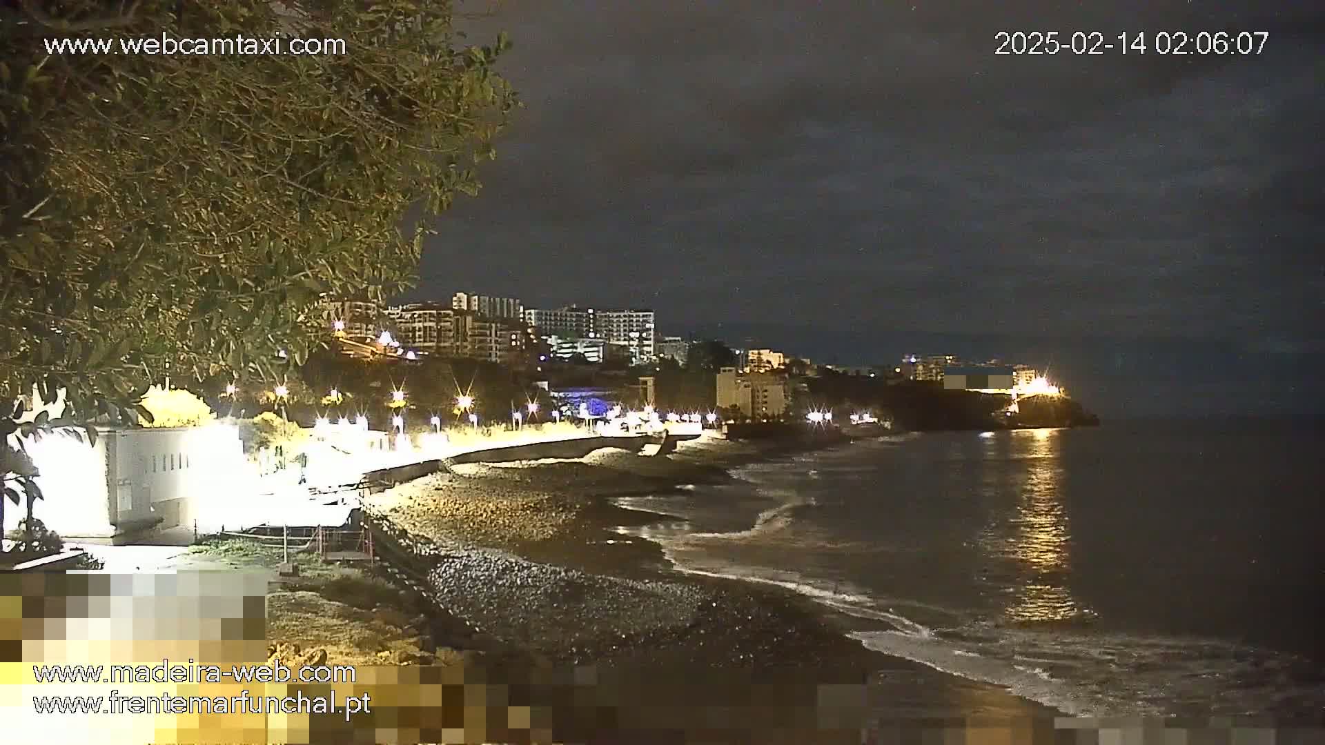 A nighttime view of a dark, pebble beach with gentle waves lapping the shore, under a cloudy sky, with illuminated buildings and streetlights visible along the coastline.