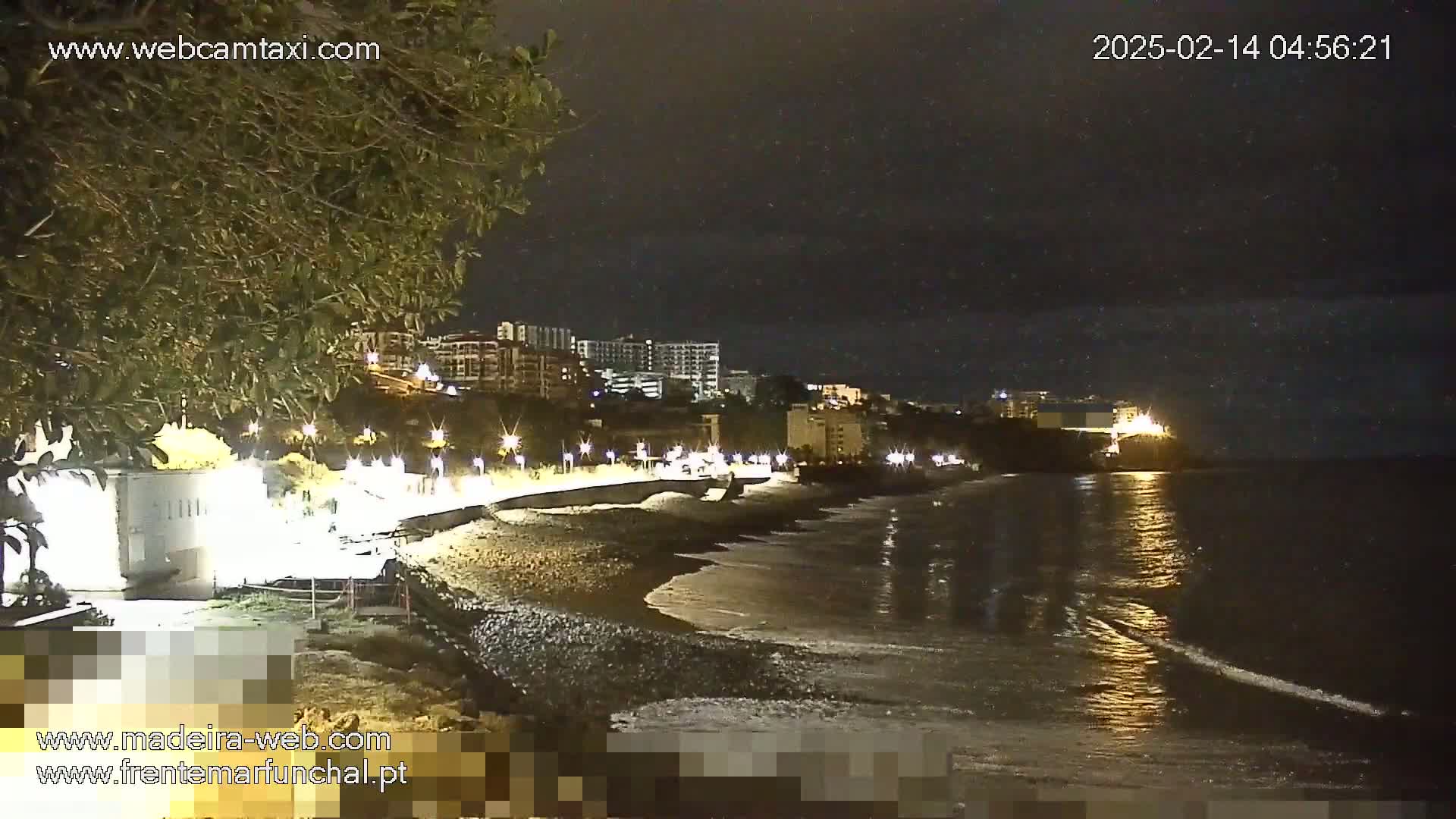 A dark, nighttime scene shows a beach with gentle waves lapping the shore, illuminated buildings in the background, and a few lights along a paved walkway.