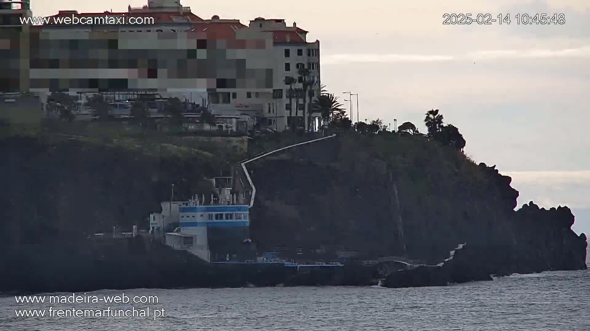 A multi-story building sits atop a dark cliff overlooking a calm body of water under an overcast sky.