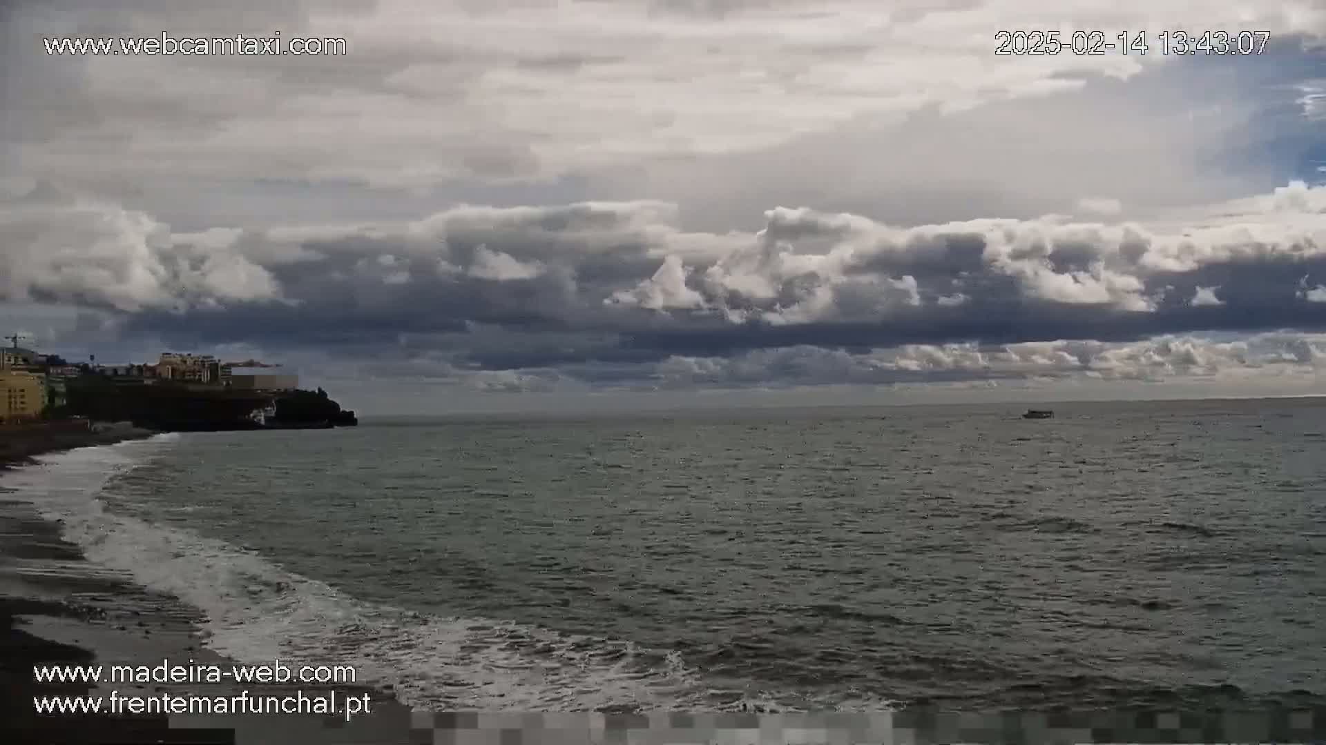 A dark, cloudy sky hangs over a dark-grey ocean with small waves lapping a dark-sand beach near a town.