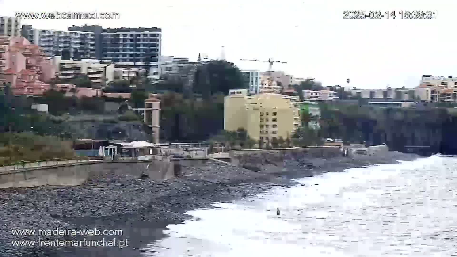 A rocky shoreline with moderate waves laps against a seawall in front of buildings on an overcast day.