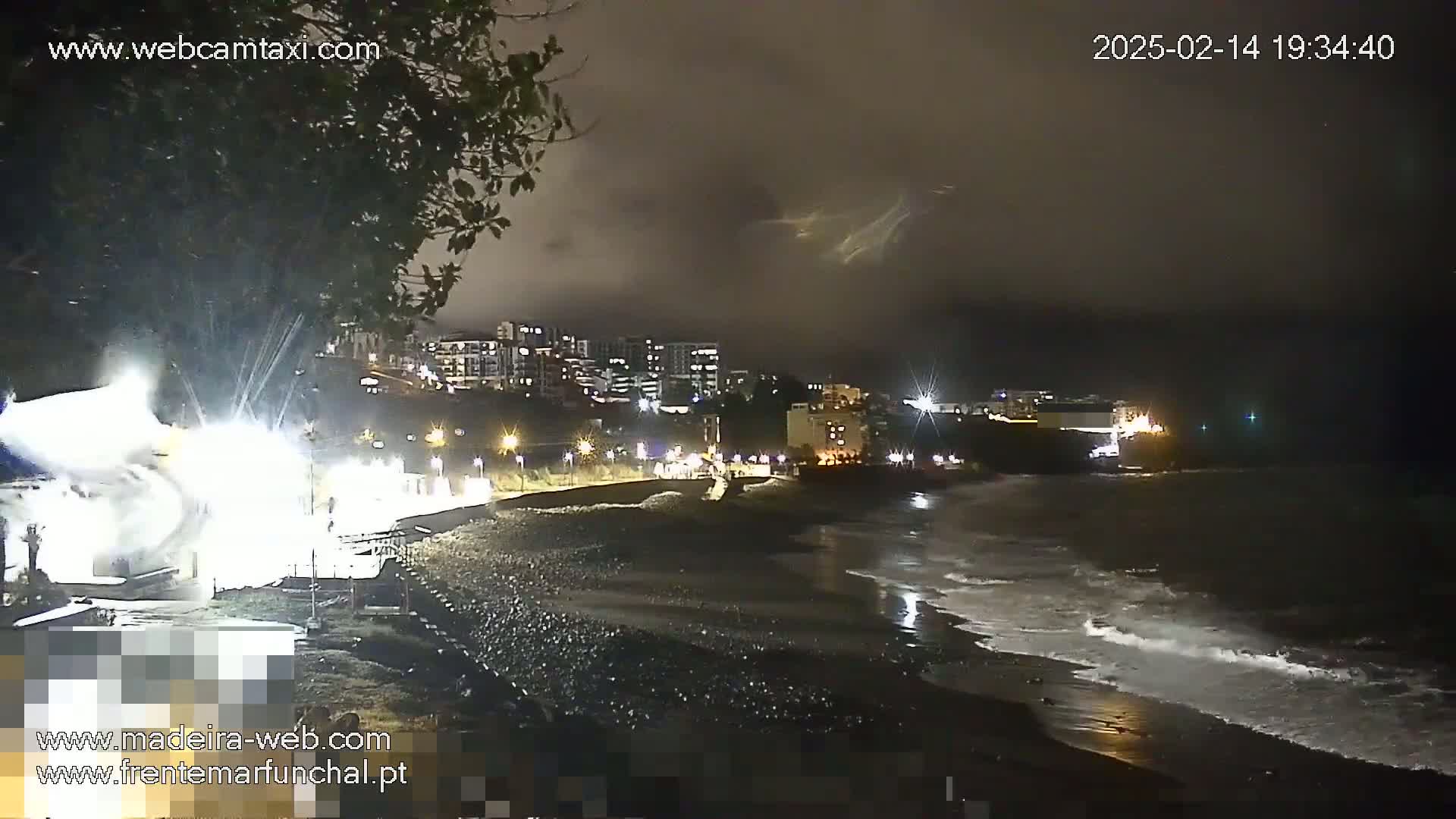 A nighttime view of a dark, rocky beach with gentle waves under a cloudy sky, showing a town's lights in the background.