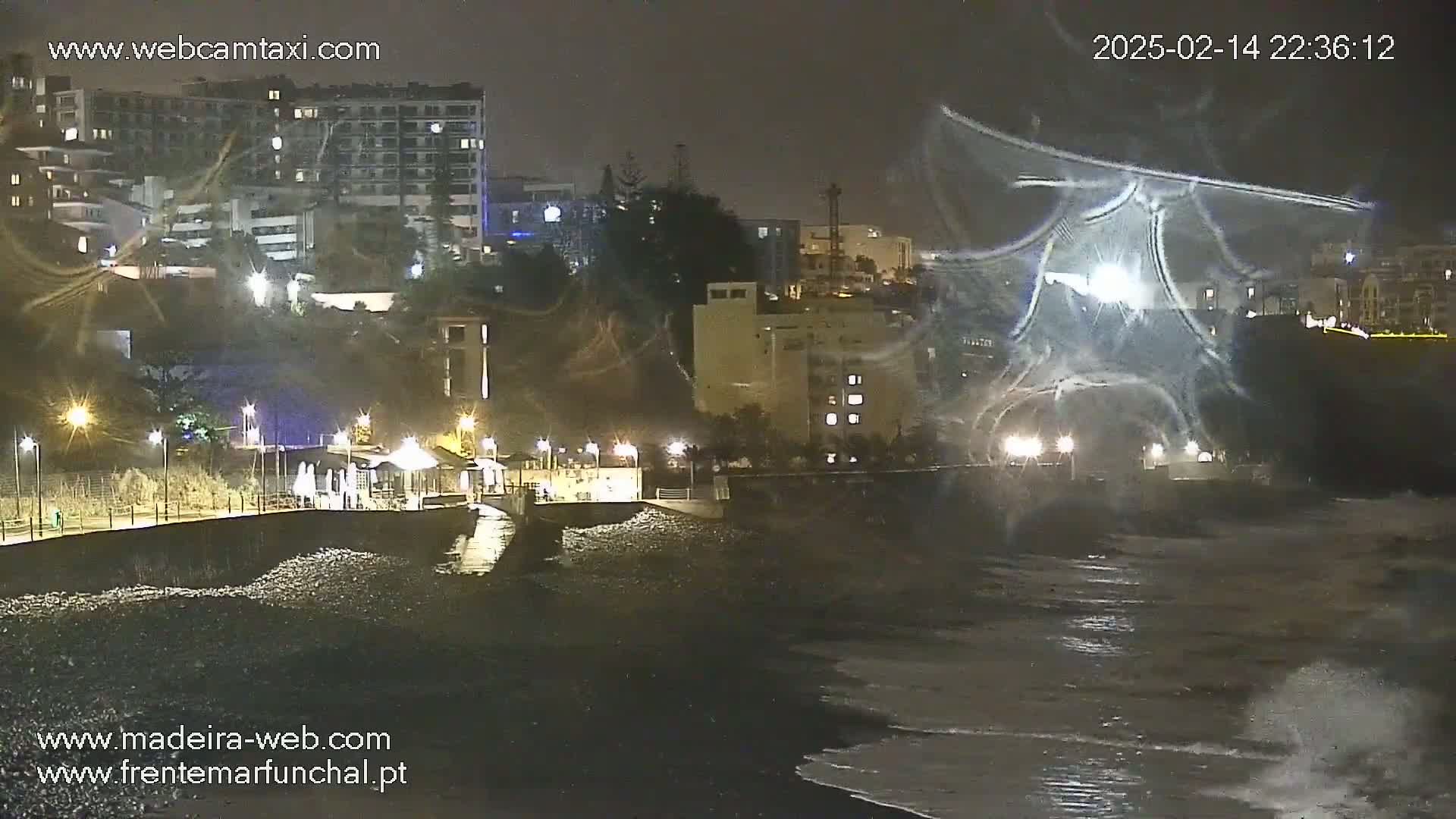 A nighttime coastal scene shows a dark, rocky beach with waves lapping the shore, illuminated buildings, and streetlights along a paved waterfront.