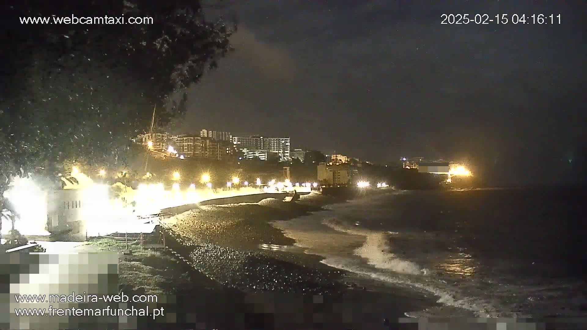 A nighttime view of a dark, rocky beach with gentle waves lapping the shore, illuminated buildings in the background, and a few lights lining the waterfront.