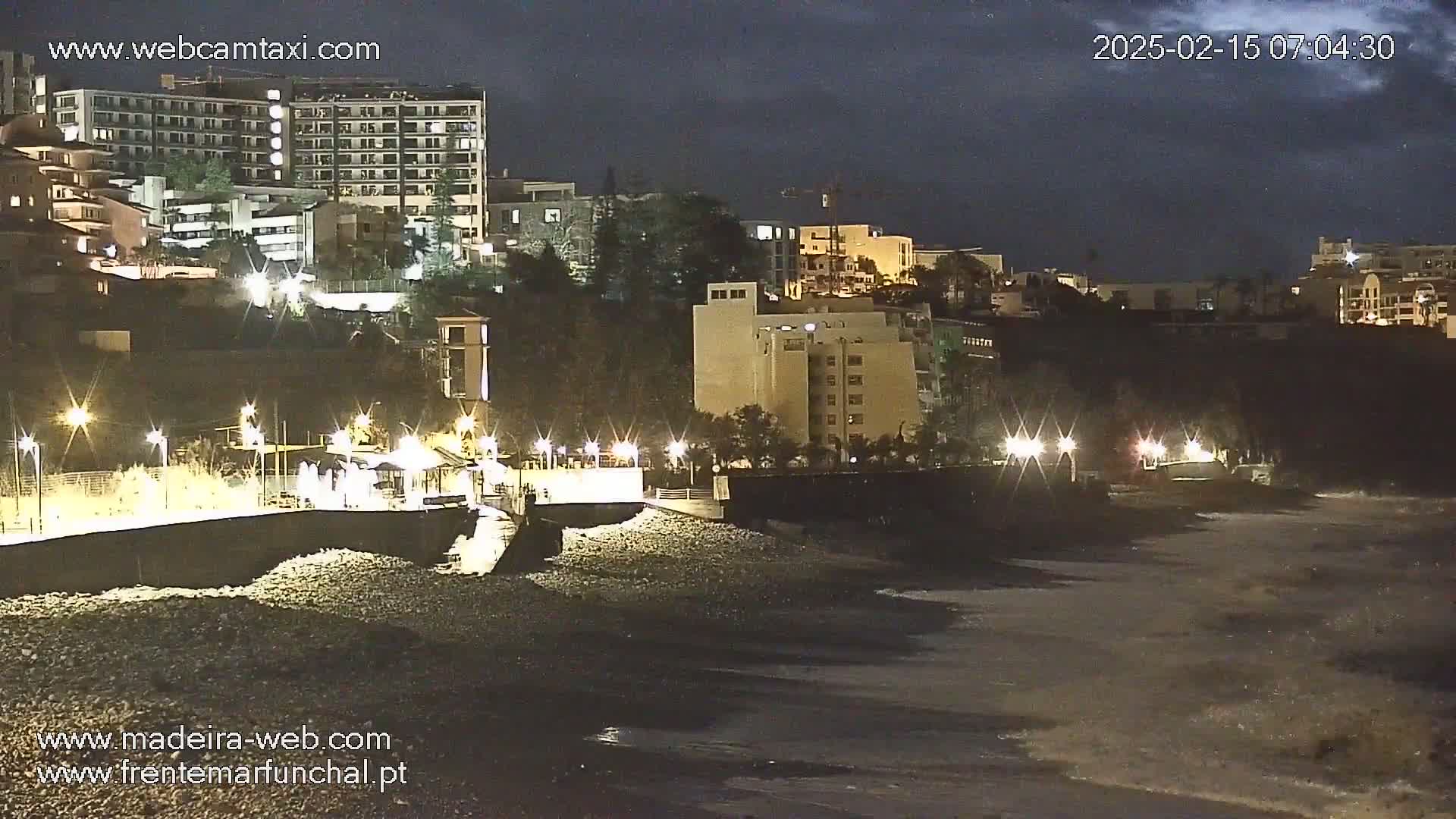 At night, under a dark sky, a rocky shoreline is illuminated by lights lining a paved walkway that leads to several buildings perched on a hill overlooking the ocean.