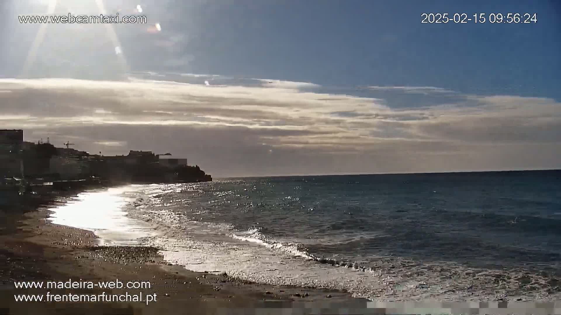 A sandy beach with gentle waves laps the shore under a partly cloudy sky, the sun shining brightly.