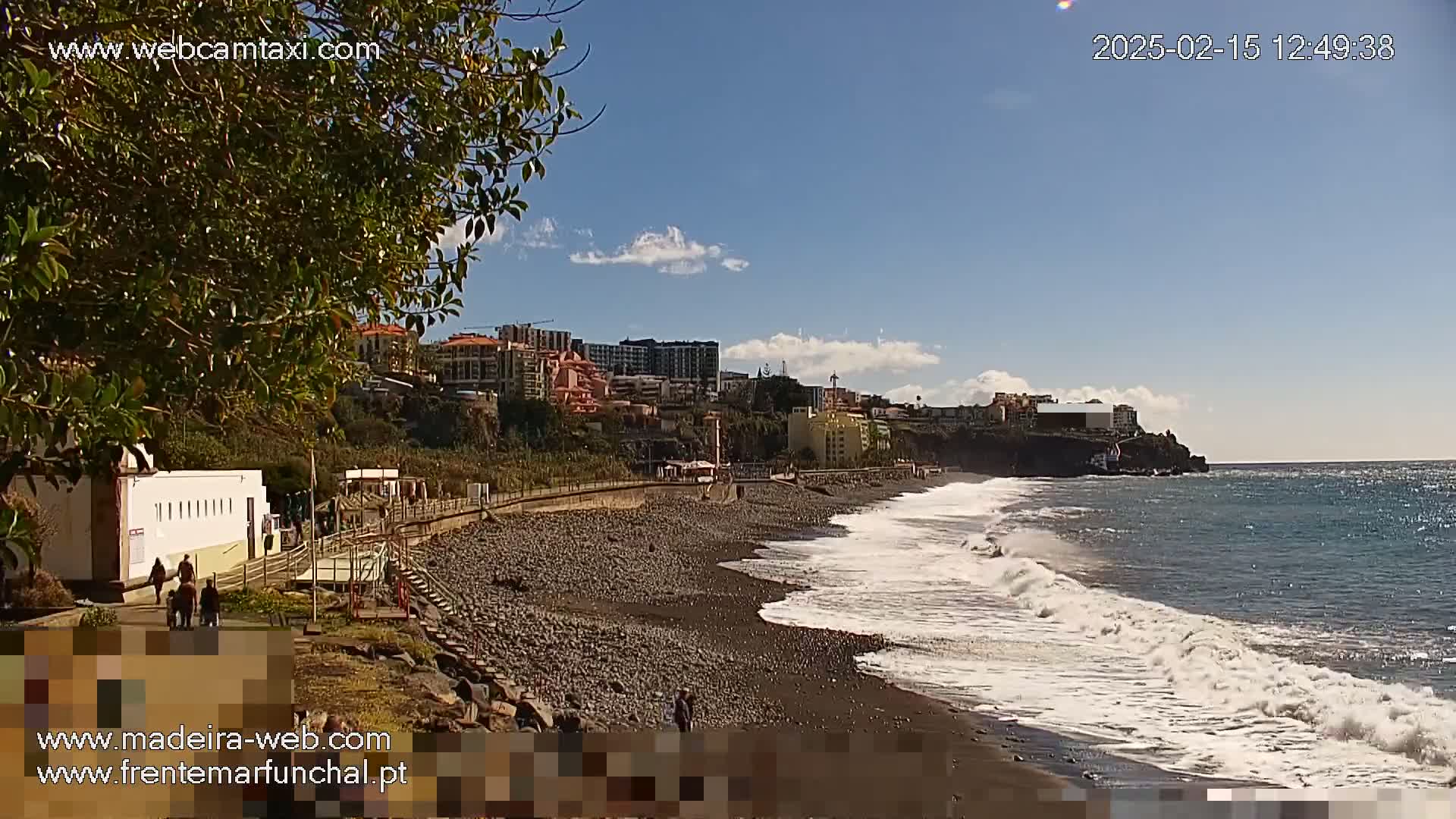 A dark-sand beach with moderate waves under a mostly sunny sky features buildings and vegetation along a paved walkway behind it.