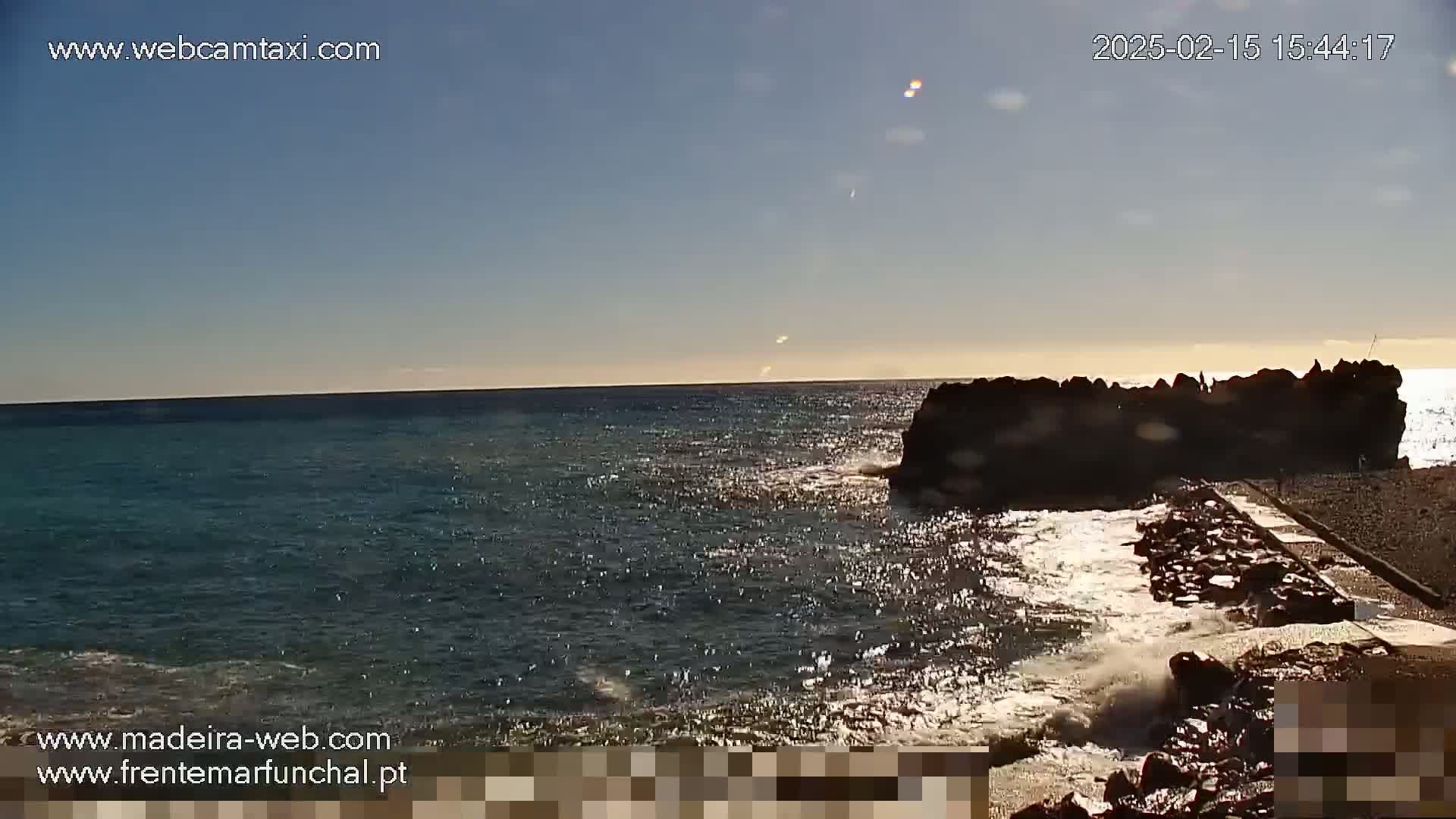 A rocky outcropping juts into a calm, sunlit ocean under a clear sky.