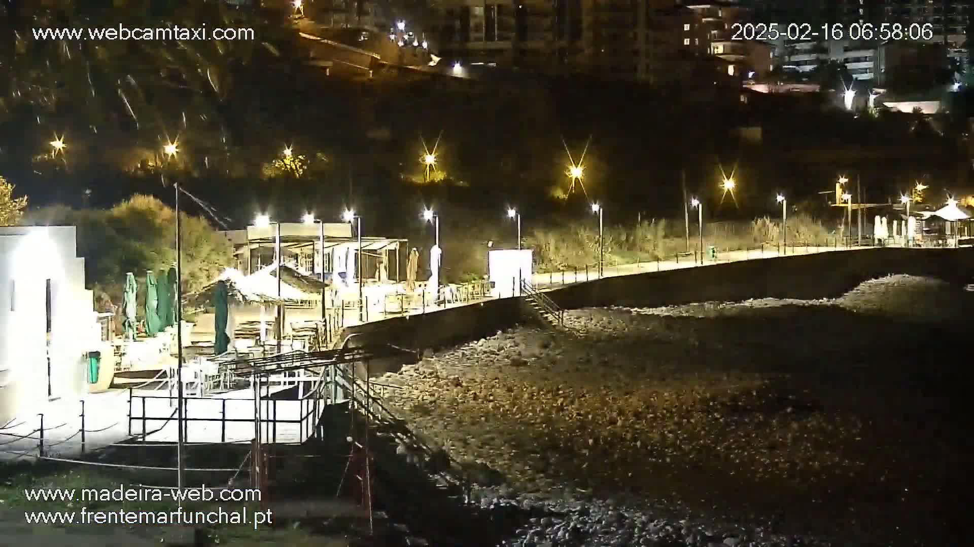 A nighttime view of a rocky beach with illuminated buildings and a boardwalk along the shore.