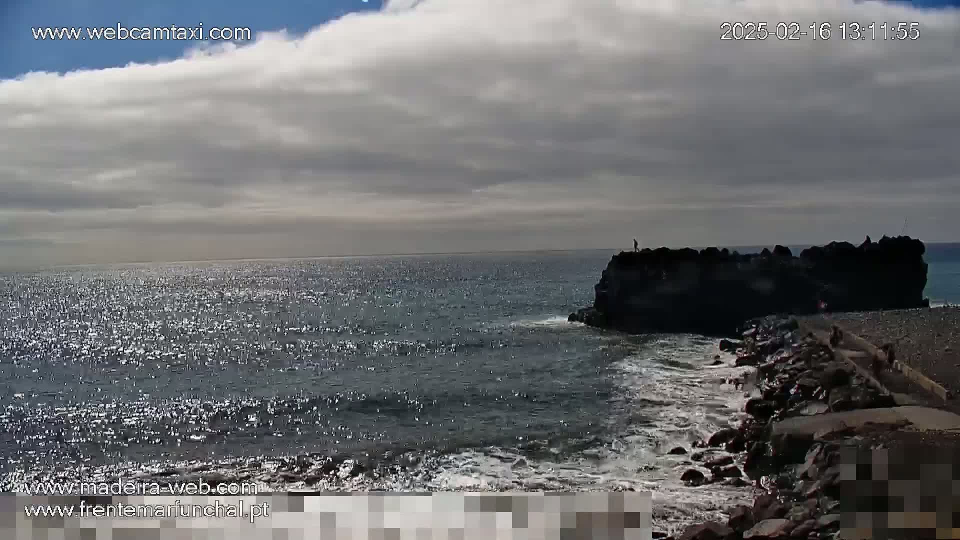 A partly cloudy day shows a rocky shoreline with a person standing on a rock outcropping in the ocean; gentle waves lap against the shore.
