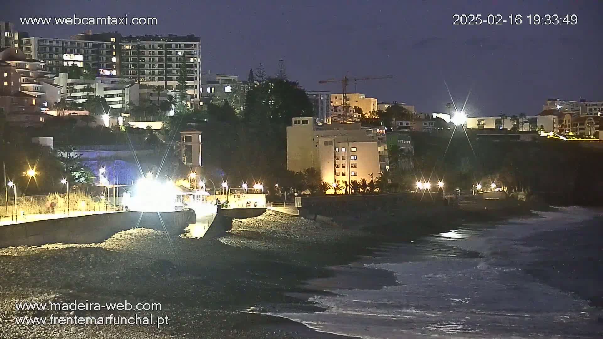 A nighttime view of a dark, rocky beach with gentle waves lapping the shore, illuminated by streetlights, with buildings and a crane visible on the hillside above.