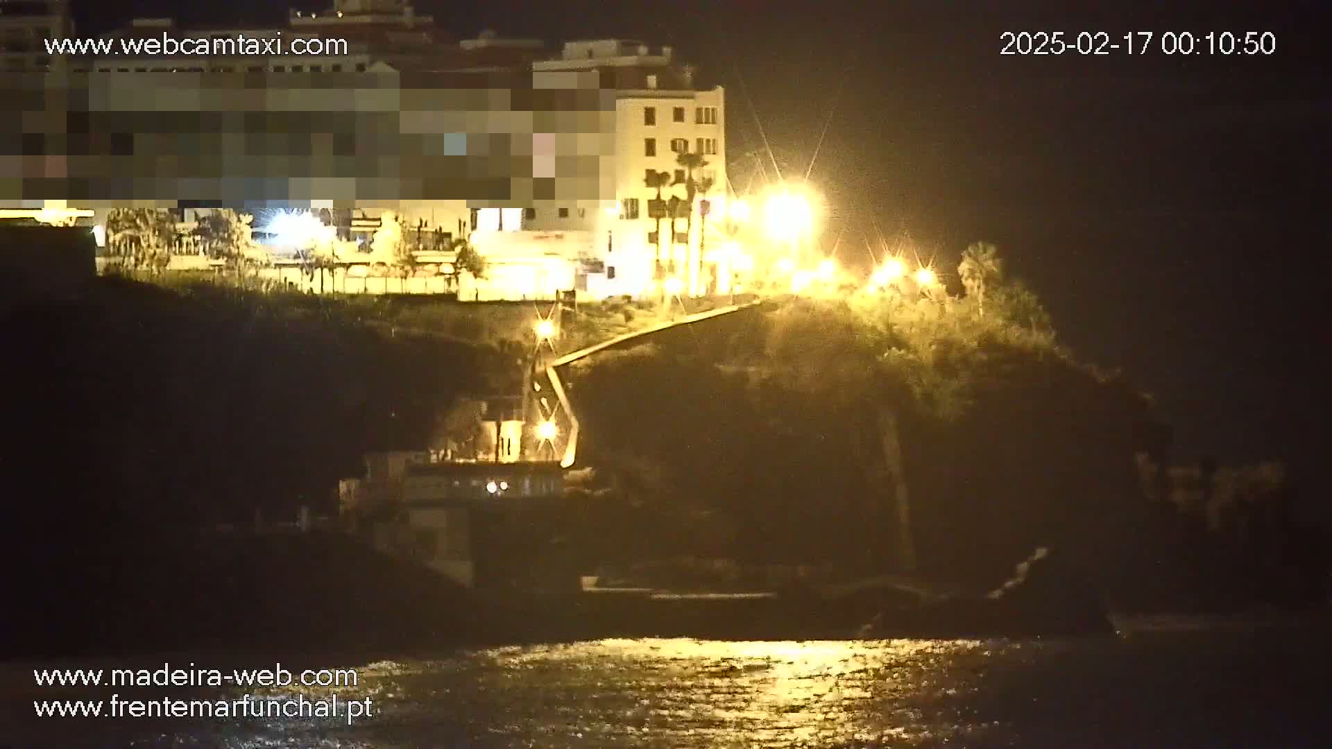 A nighttime coastal scene shows a building illuminated by streetlights overlooking a dark, calm body of water.