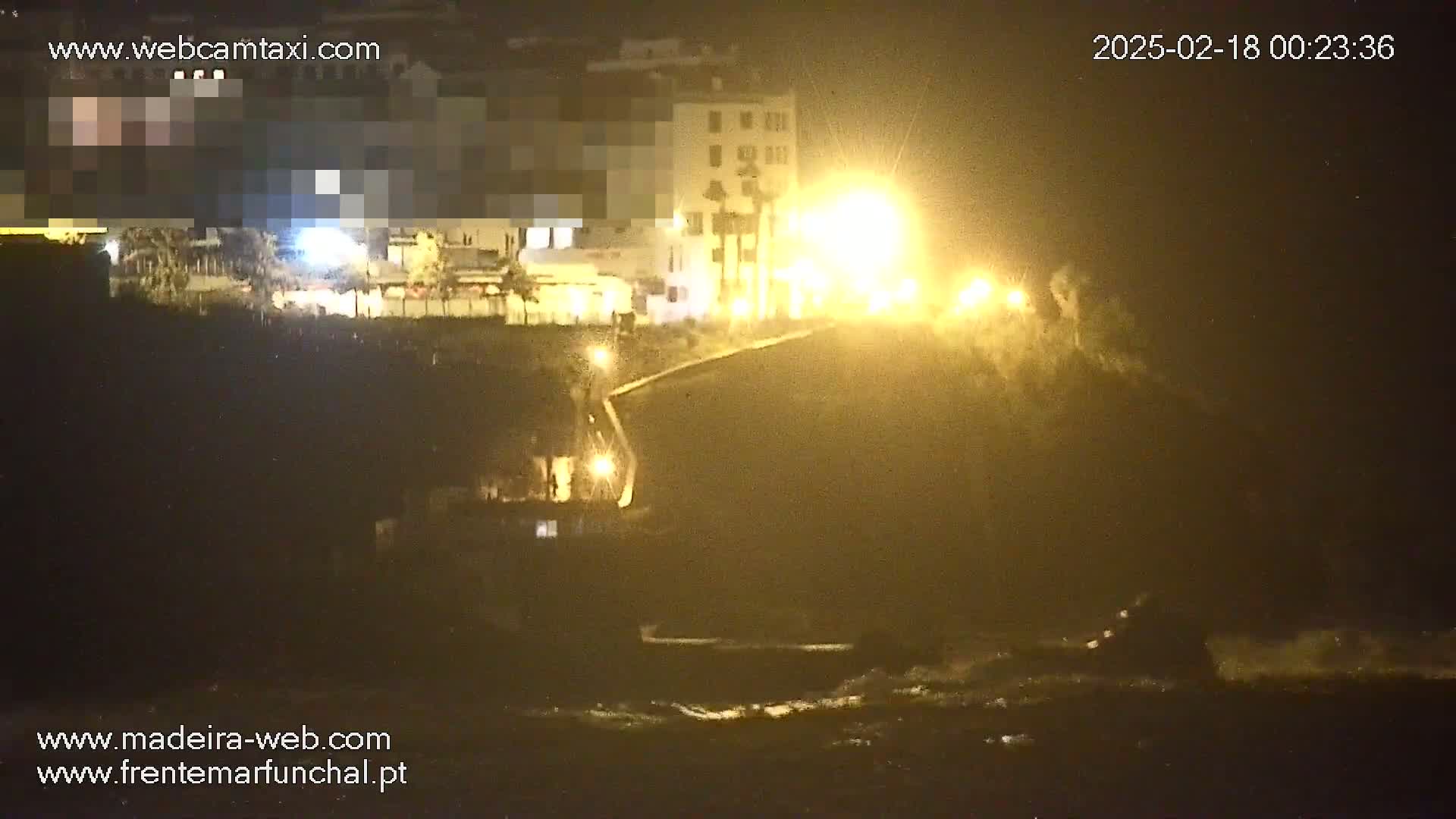 A nighttime coastal scene shows brightly lit buildings overlooking a dark, wavy ocean under hazy, yellow-tinged conditions.