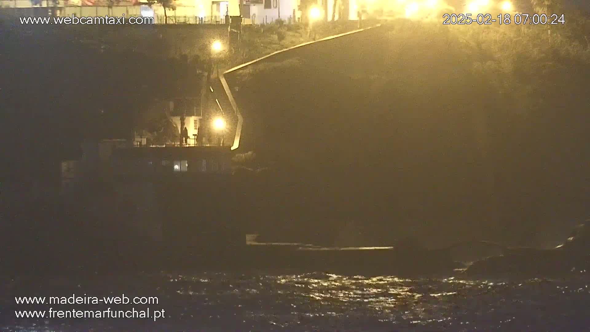 A dark, nighttime coastal scene shows a structure extending out over choppy water, illuminated by several lights against a hazy, possibly foggy, sky.